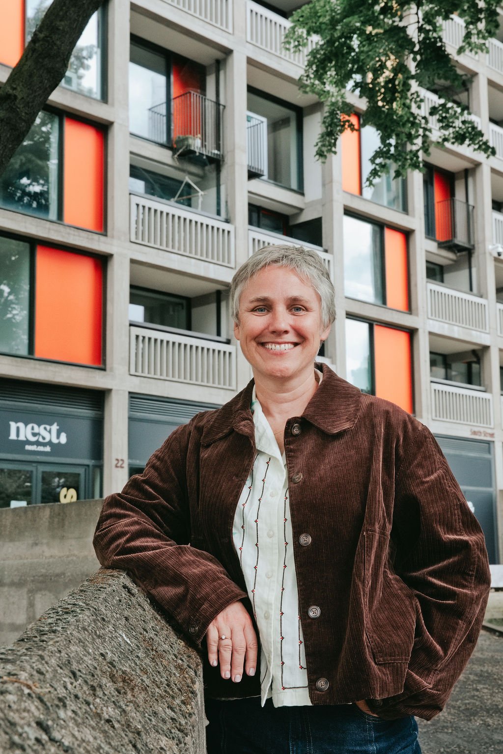 Smiling woman with short gray hair wearing a brown jacket and white shirt, leaning on a concrete barrier in front of a modern building with orange accents and balconies.
