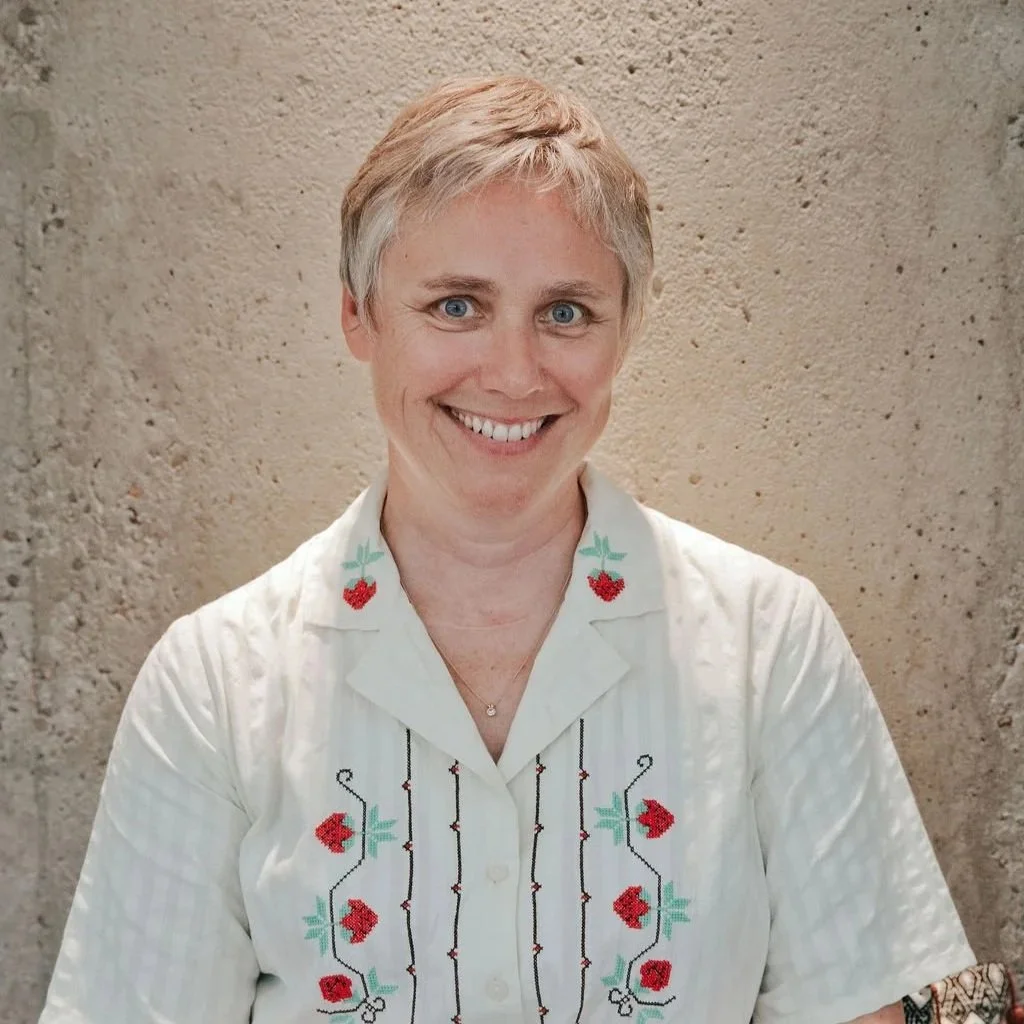 A woman with short blonde hair and blue eyes smiling at the camera, wearing a white embroidered shirt with colorful floral and strawberry patterns, standing against a textured beige wall.