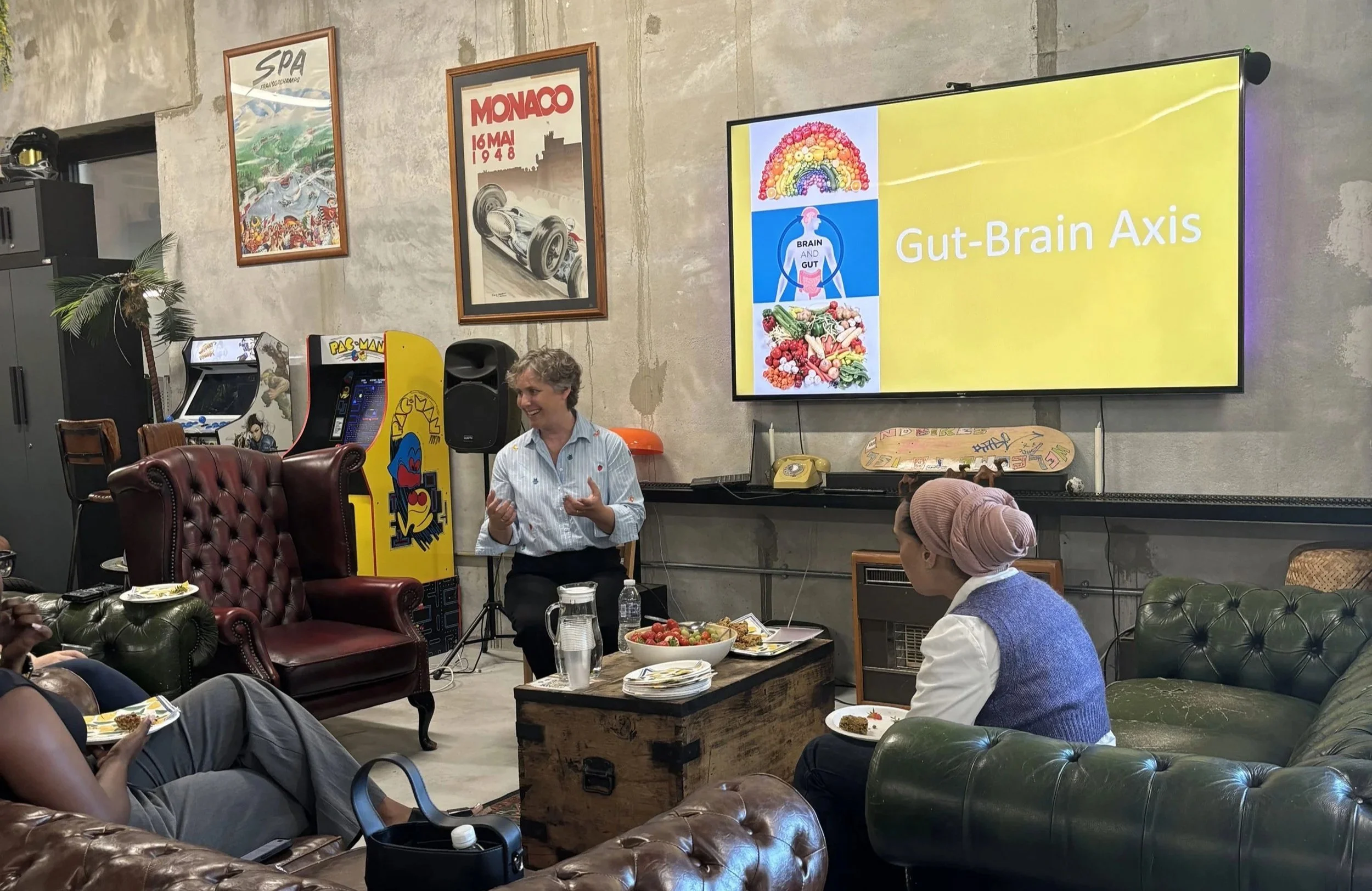 A woman with short gray hair speaking in front of a group at a casual gathering. There is a large screen displaying a presentation titled "Gut-Brain Axis" with images of food and a diagram of the brain and gut. The room has vintage arcade machines, framed posters including one of a Monaco race car, and various chairs and tables with snacks and drinks.