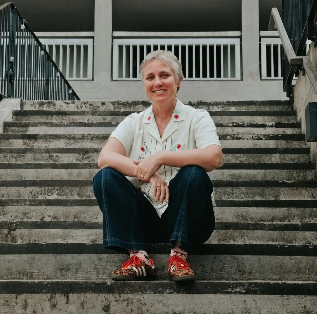 A woman sits on outdoor concrete stairs, smiling at the camera, with a modern building in the background.