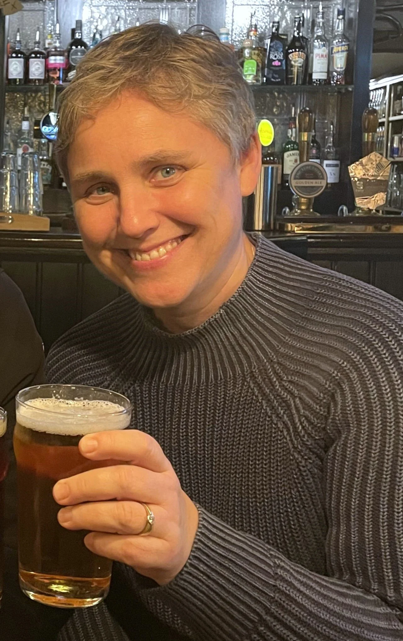 A smiling man with light brown hair in a striped sweater holding a glass of beer in a bar setting.