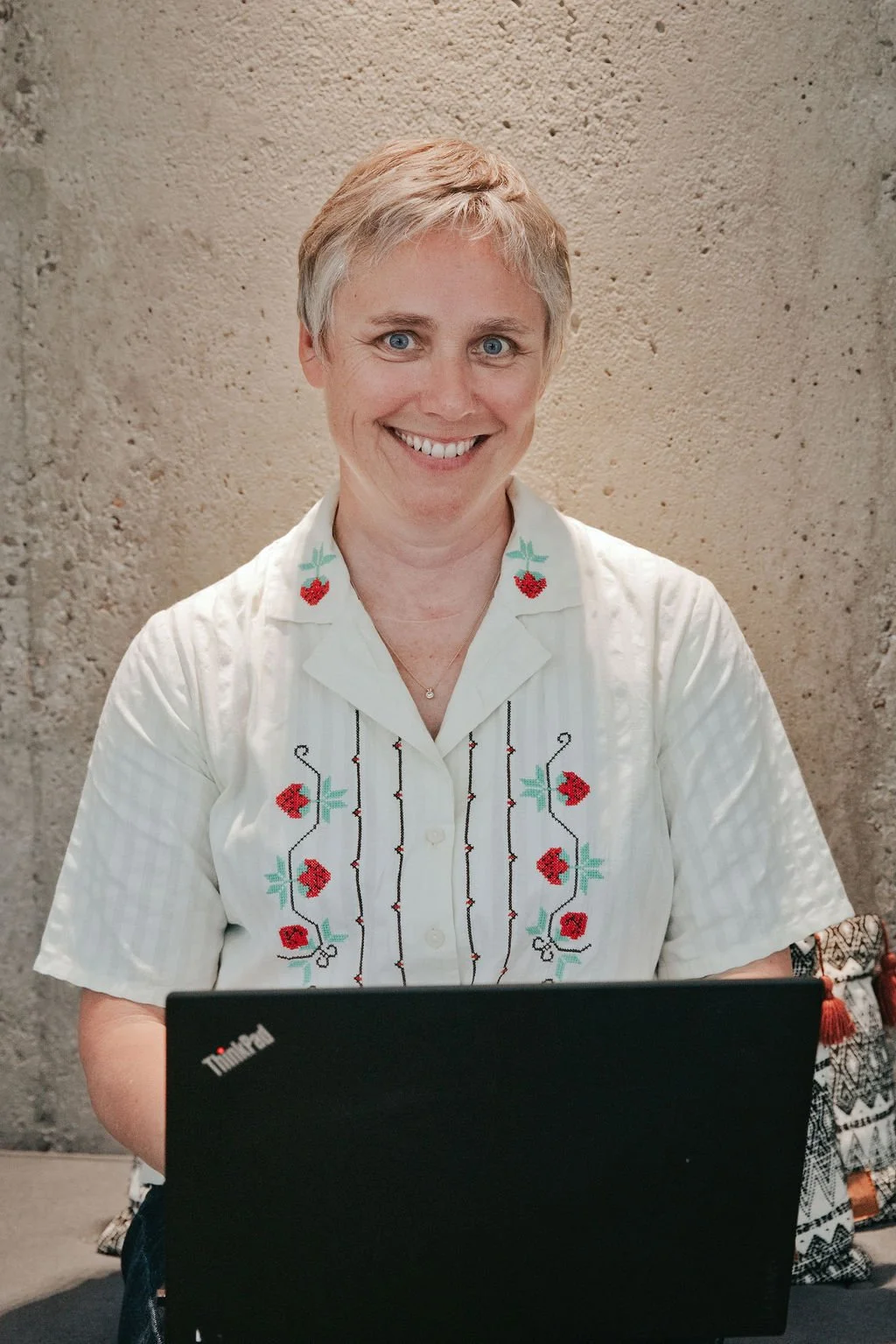 A woman with short, blond hair smiling at the camera, wearing a white embroidered shirt with red and teal floral patterns, sitting in front of a textured beige wall, with a black laptop and a colorful bag nearby.