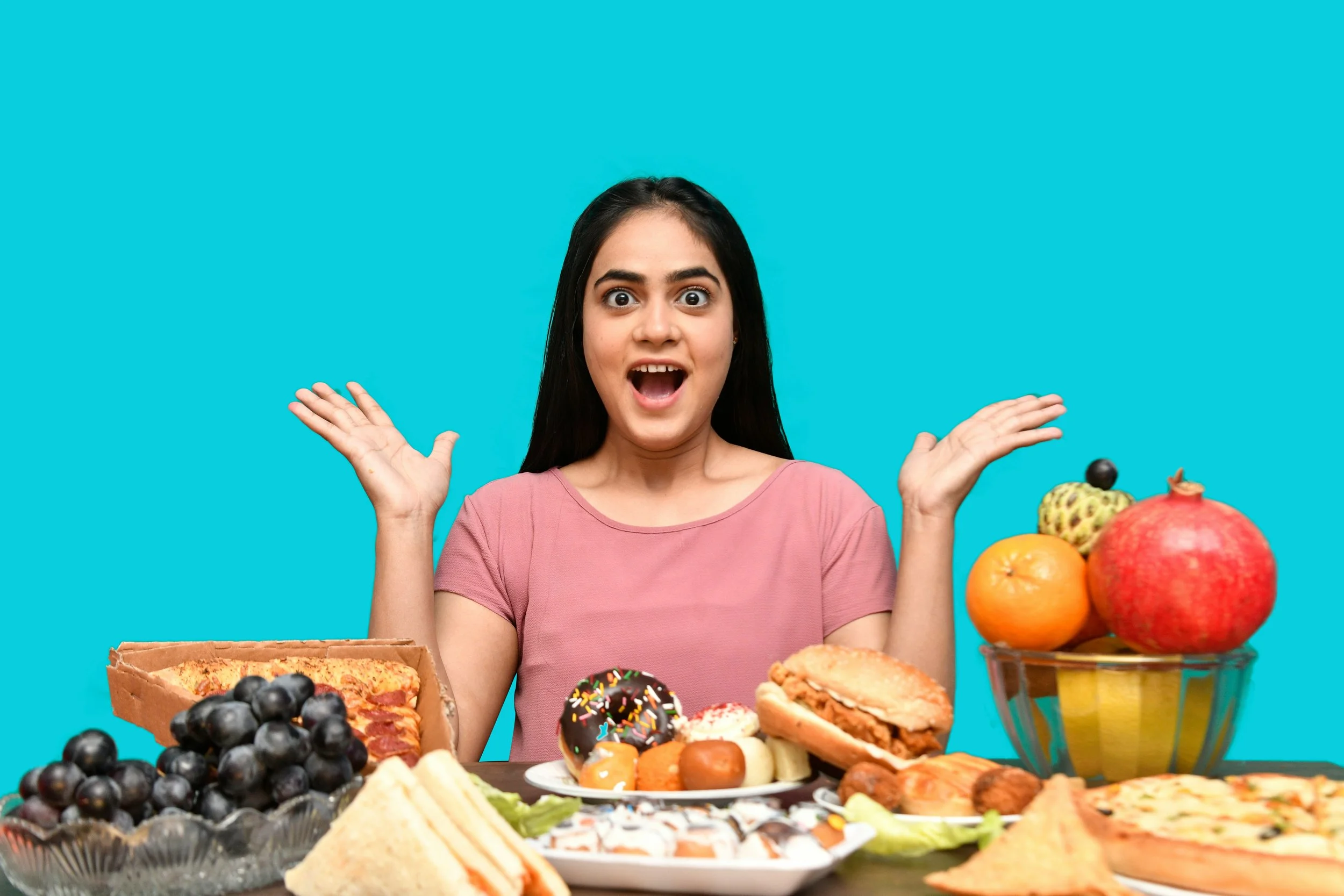 A young woman with dark hair and a pink shirt, looking surprised with her mouth open and hands near her face, seated at a table filled with various desserts and fruits against a bright blue background.