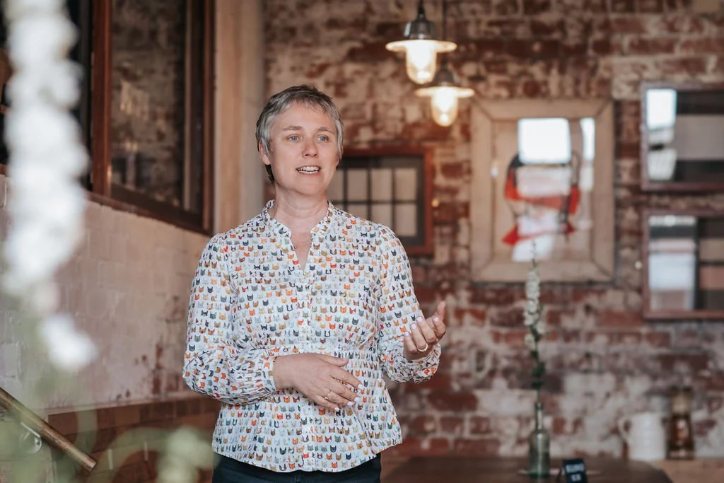 A woman with short gray hair speaking indoors, wearing a white blouse with colorful cat print, in a rustic brick-walled space with hanging lights and framed artwork.