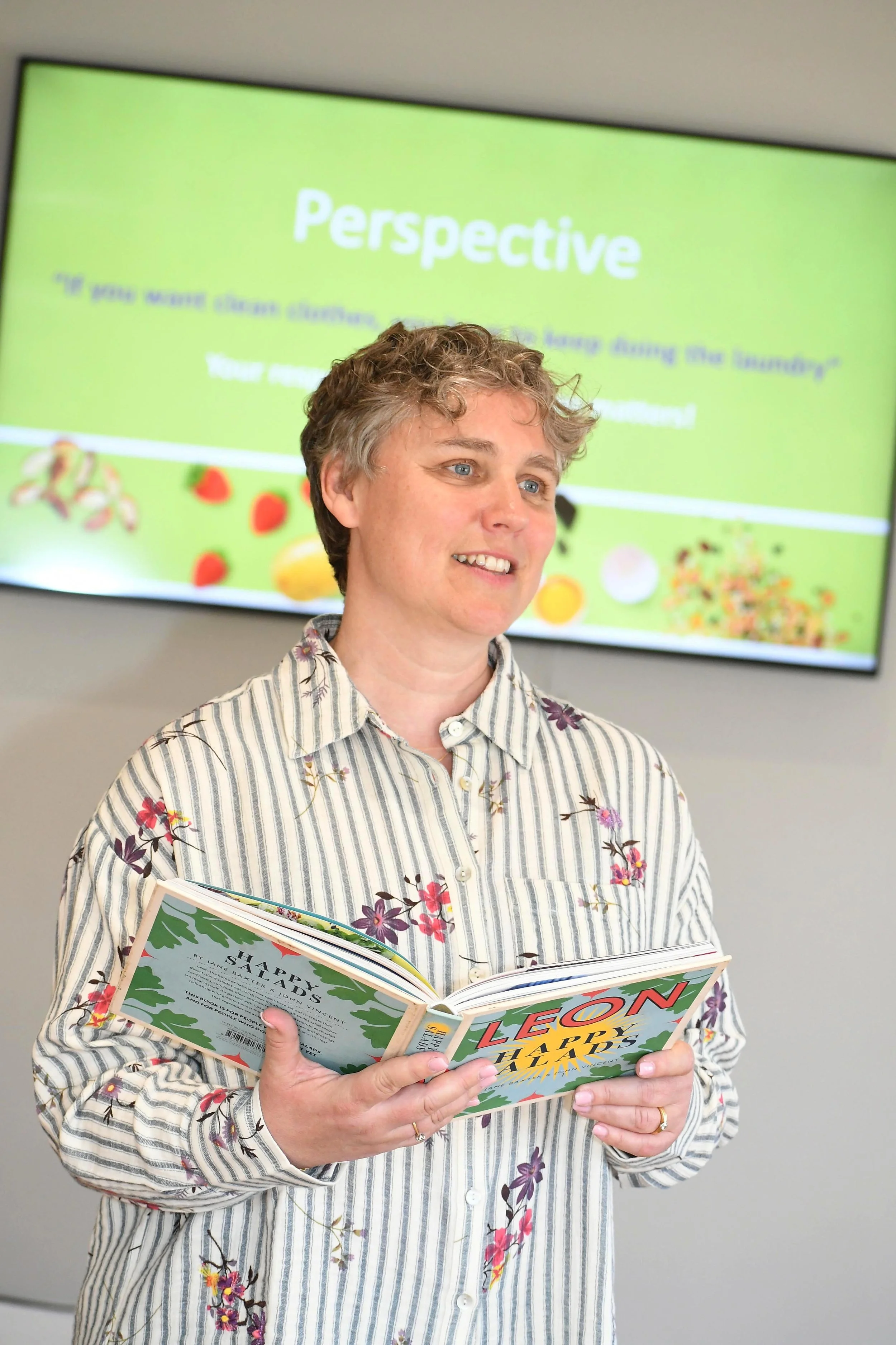 A woman with short, curly hair, wearing a striped floral shirt, stands in front of a presentation screen, holding an open book titled 'LEON Happy Holidays'.