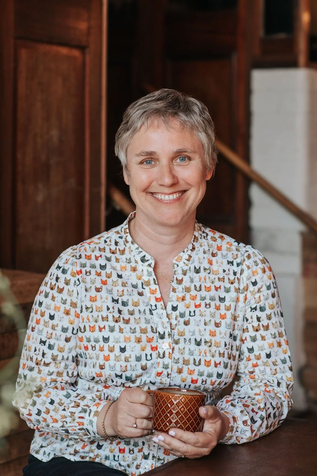 A smiling woman with short gray hair holding a brown patterned mug, sitting indoors with wooden walls and stairs in the background, wearing a white blouse with a colorful cat face print.