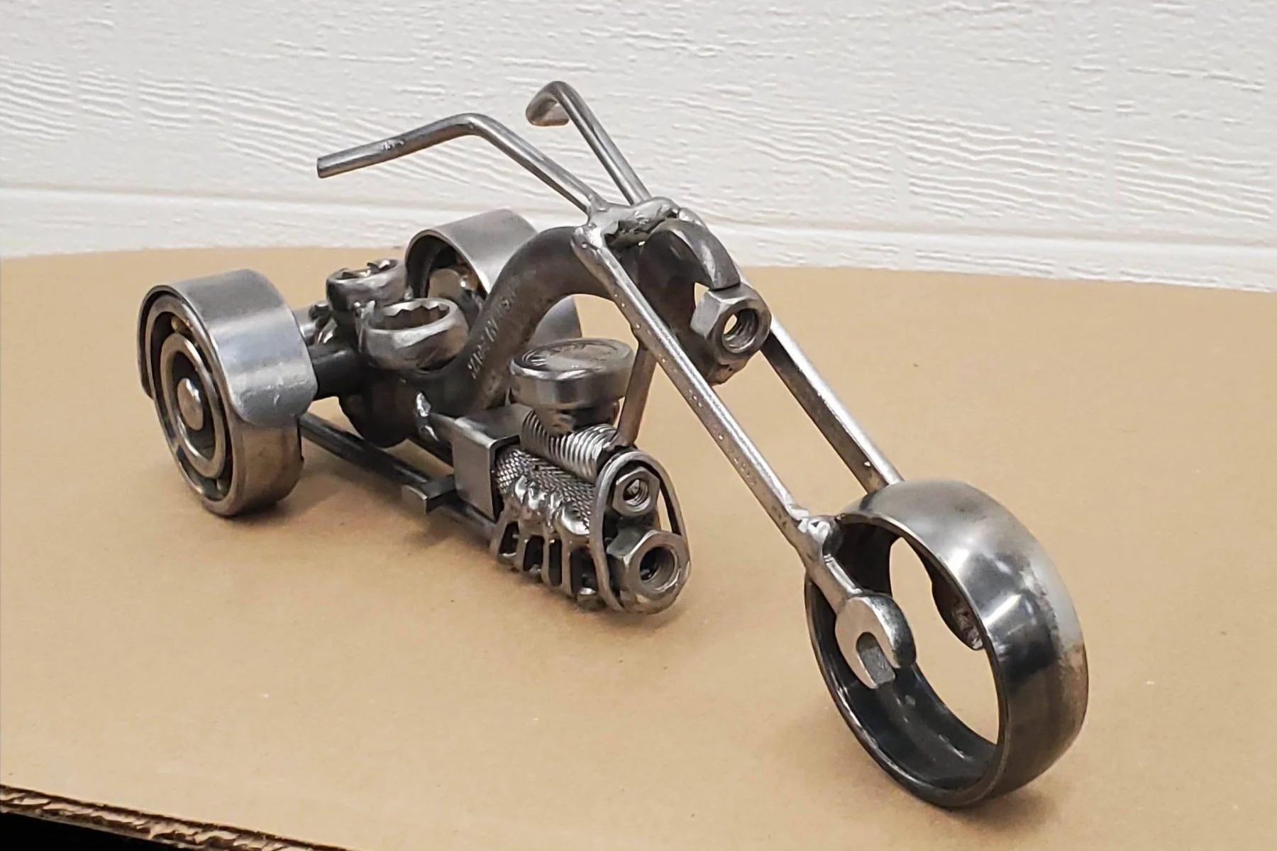 A metal sculpture of a motorcycle, made from various metal parts and scrap, placed on a beige surface against a white textured wall background.