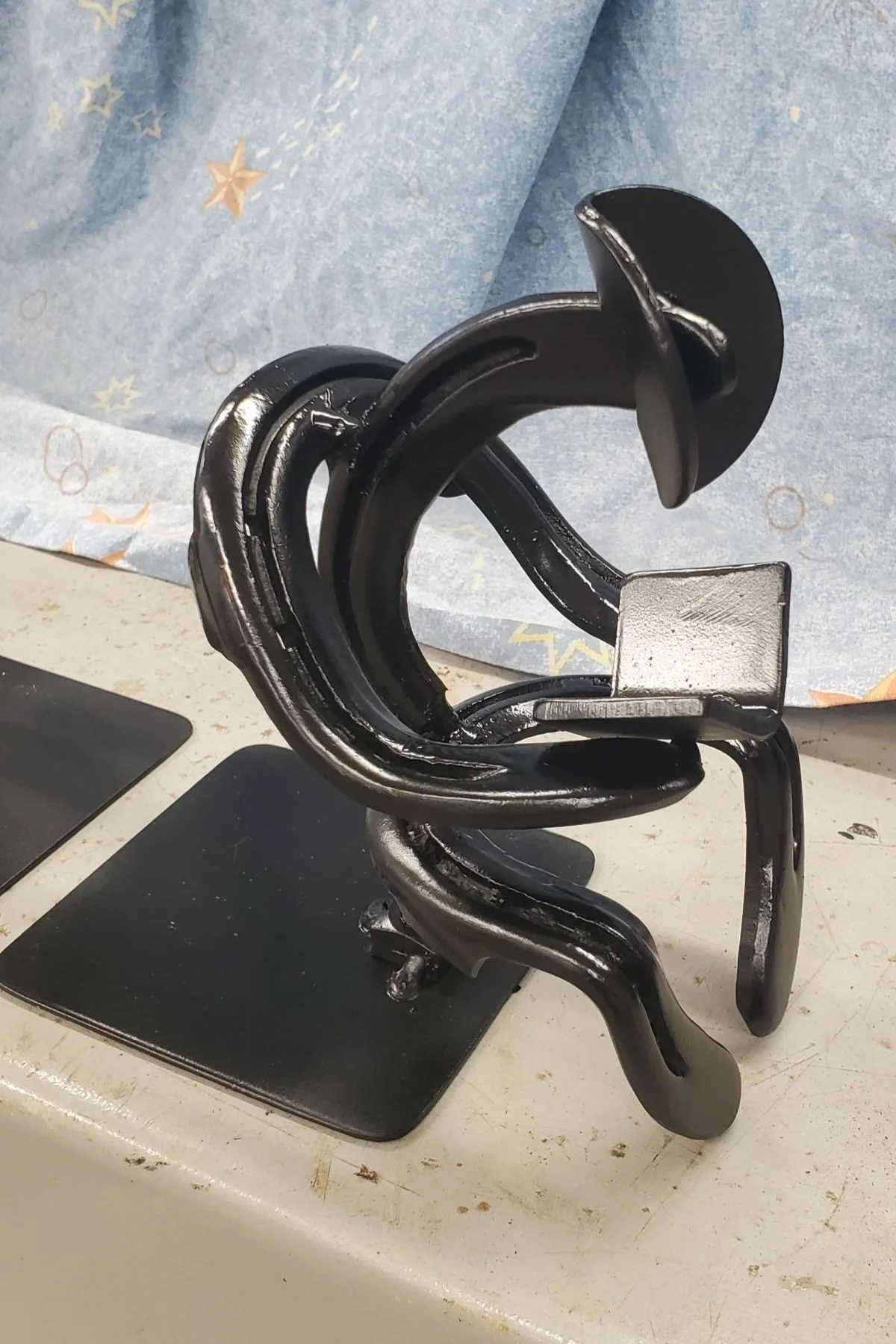 A metallic sculpture resembling a person reading a book, made of interconnected black and silver metal pieces, resting on a black square base on a white surface with a background of patterned blue paper.