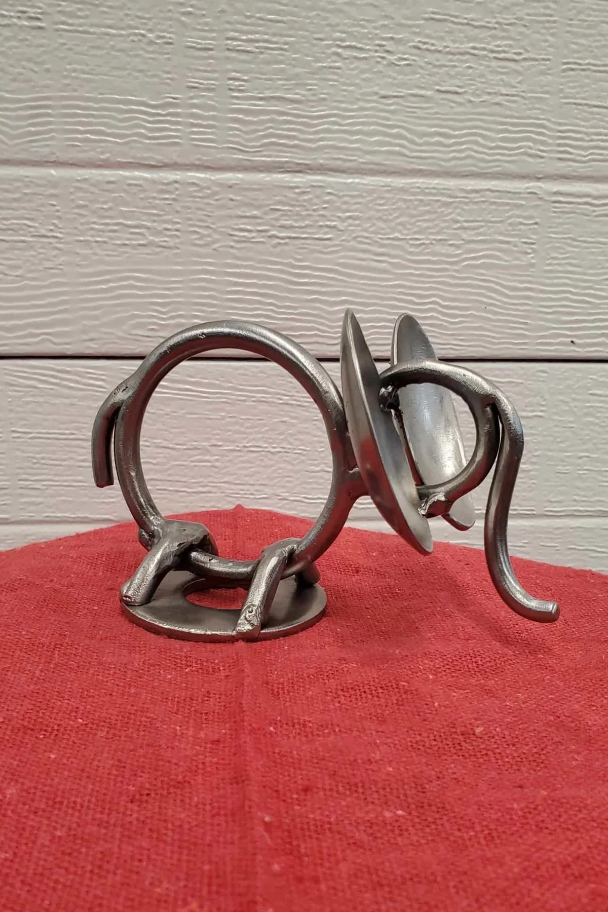 A metal elephant figurine with a rounded body, four legs, a tail, and a saddle, placed on a red fabric surface against a white wooden wall background.