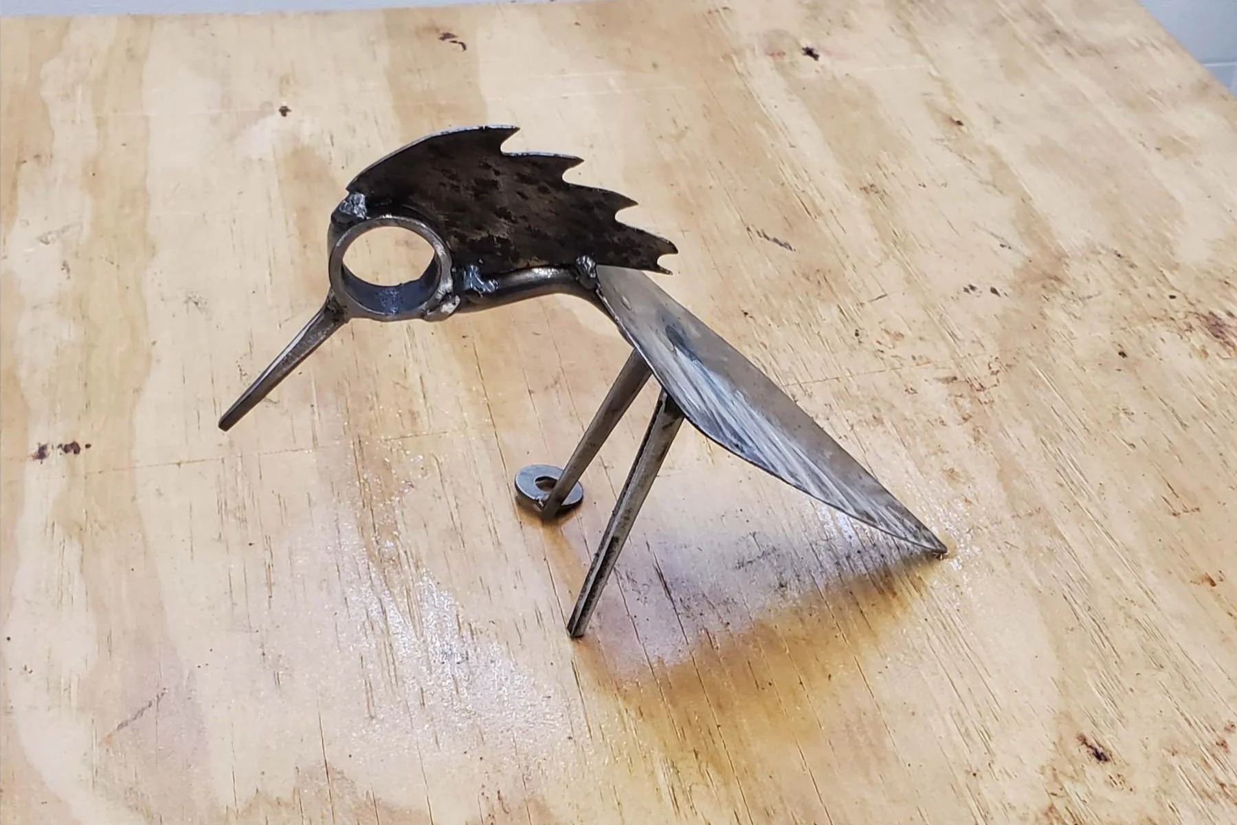 A metal sculpture of a bird, resembling a velociraptor or similar dinosaur, made from scrap metal parts, standing on a wooden surface.