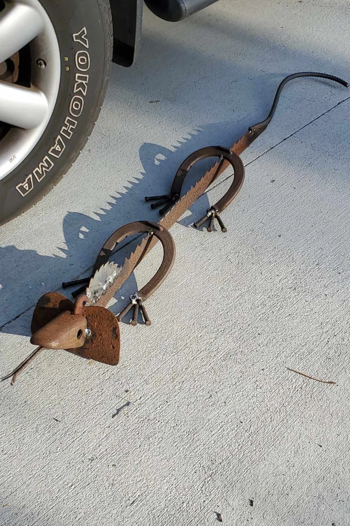 Rusty metal sculpture of an animal placed on a concrete surface near a vehicle tire.