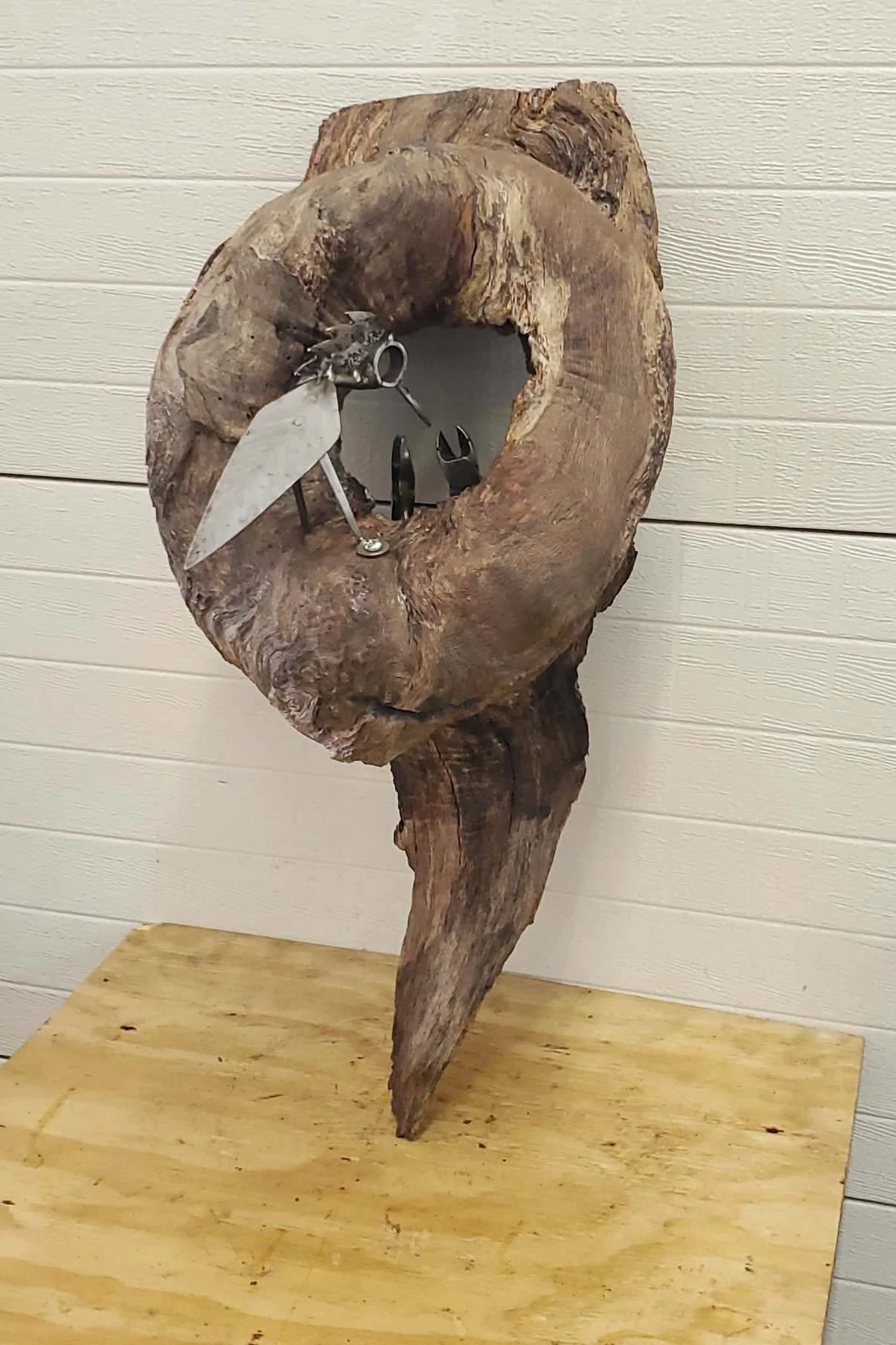 A sculpture made from scrap metal parts resembling a bird, mounted on a hollow piece of wood, against a white shiplap wall.