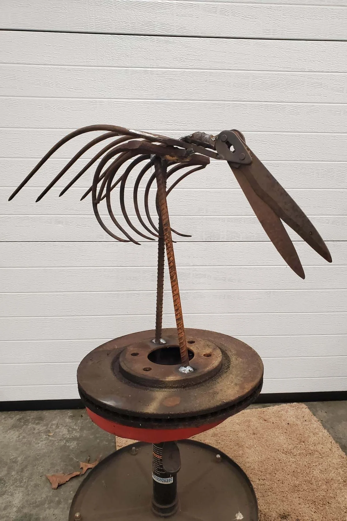 A metal sculpture resembling a hawk or bird made from reused parts, including tools and metal scraps, standing on a round base against a white textured wall.