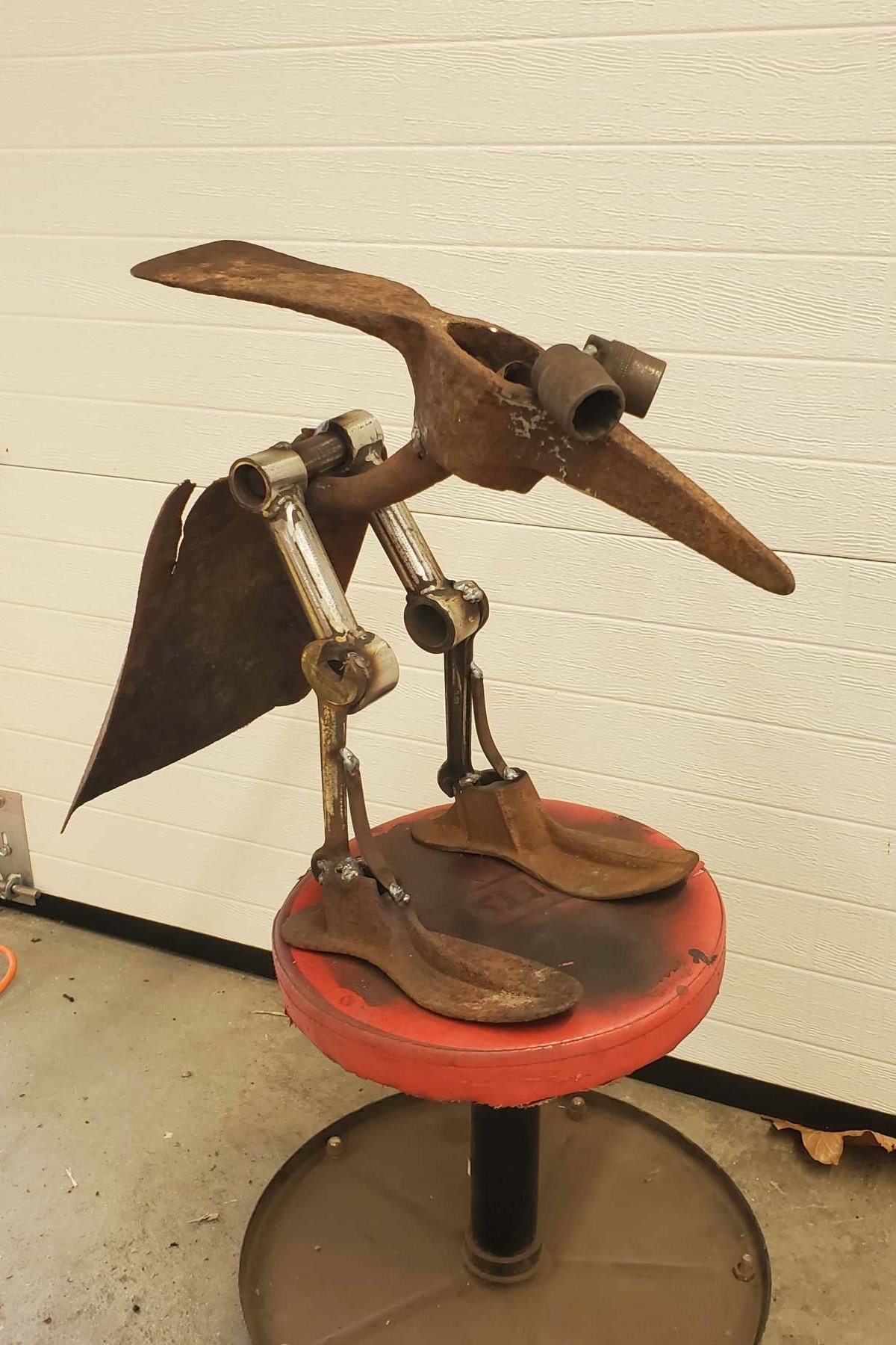 A metal sculpture of a bird with a rusted metal body, wings, and a beak. The sculpture is mounted on a red and black stool with a circular seat, set against a white wall with horizontal paneling.