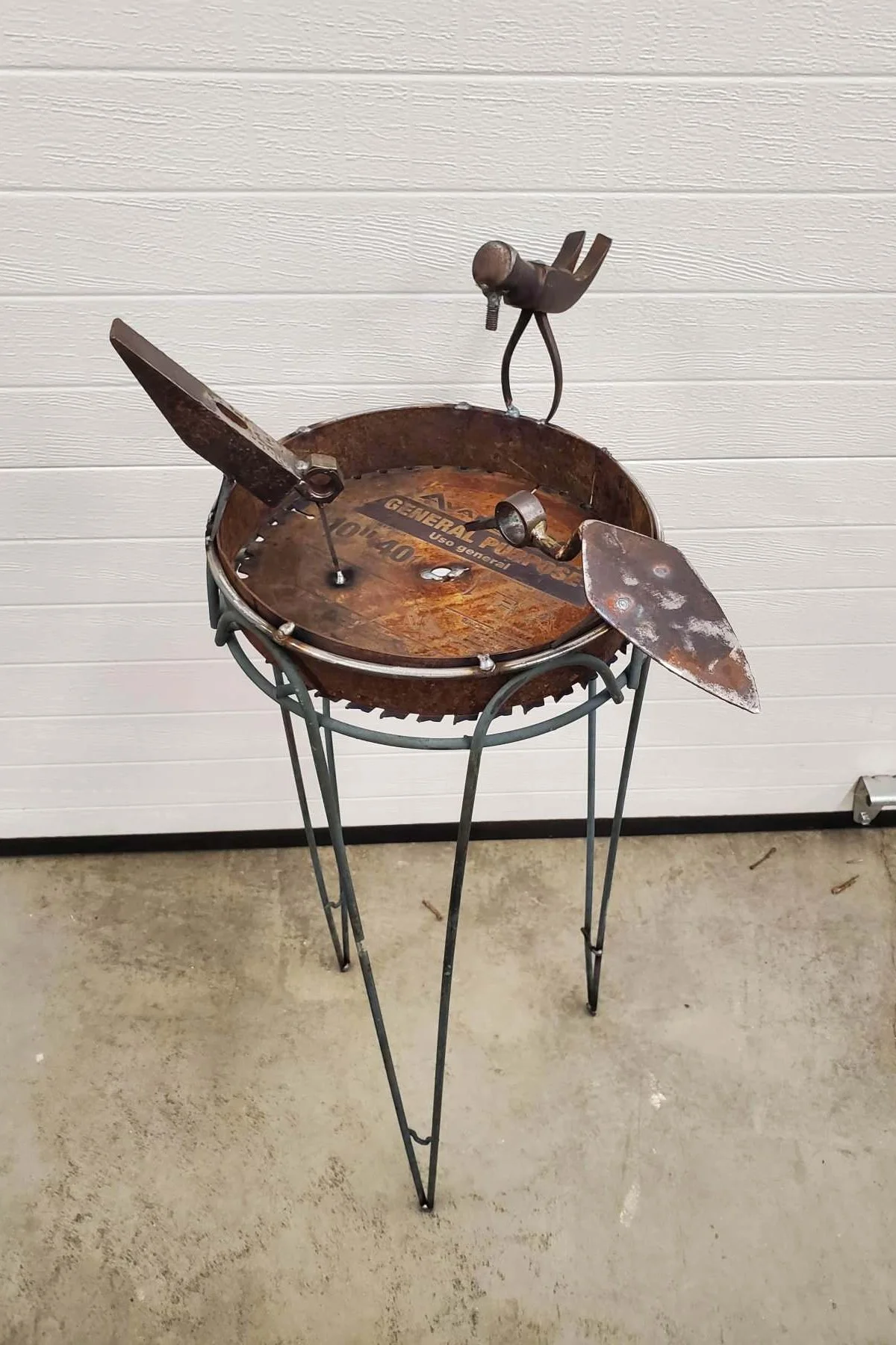 A bird sculpture made from found objects, including a rusty saw blade, a metal spoon, and various metal parts, assembled on a metal stand.