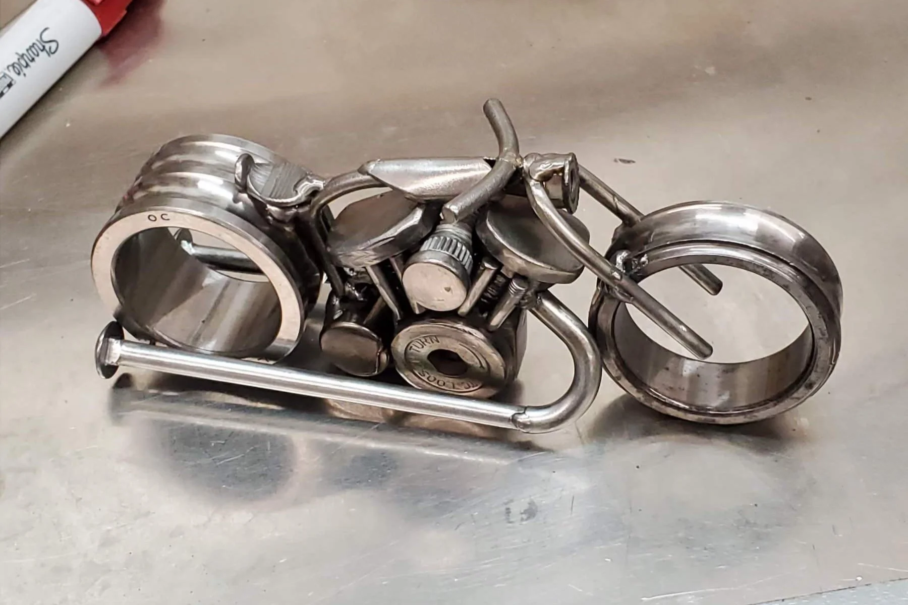 A sculpture of a motorcycle made from metallic utensils and hardware parts, placed on a metallic surface.