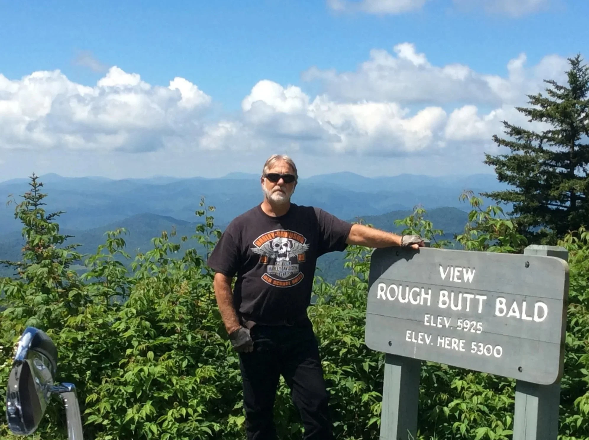 A man with gray hair, wearing sunglasses, a black Harley-Davidson T-shirt, black gloves, and black pants, stands next to a wooden sign at a mountain viewpoint. The sign reads 'View Rough Butt Bald Elev. 5925, Elev. Here 5300'. The background shows a scenic view of distant mountains under a partly cloudy sky.