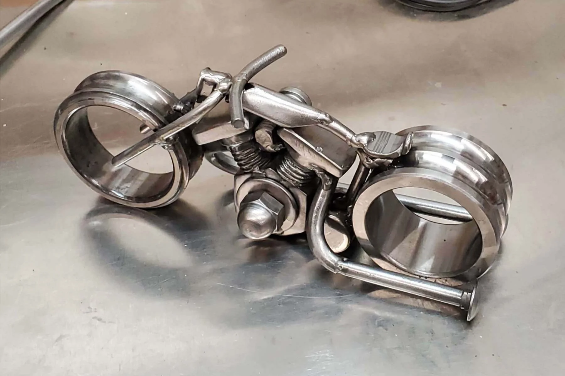 A miniature motorcycle sculpture made of metal parts, including wheels, bolts, and pipes, placed on a metallic surface.