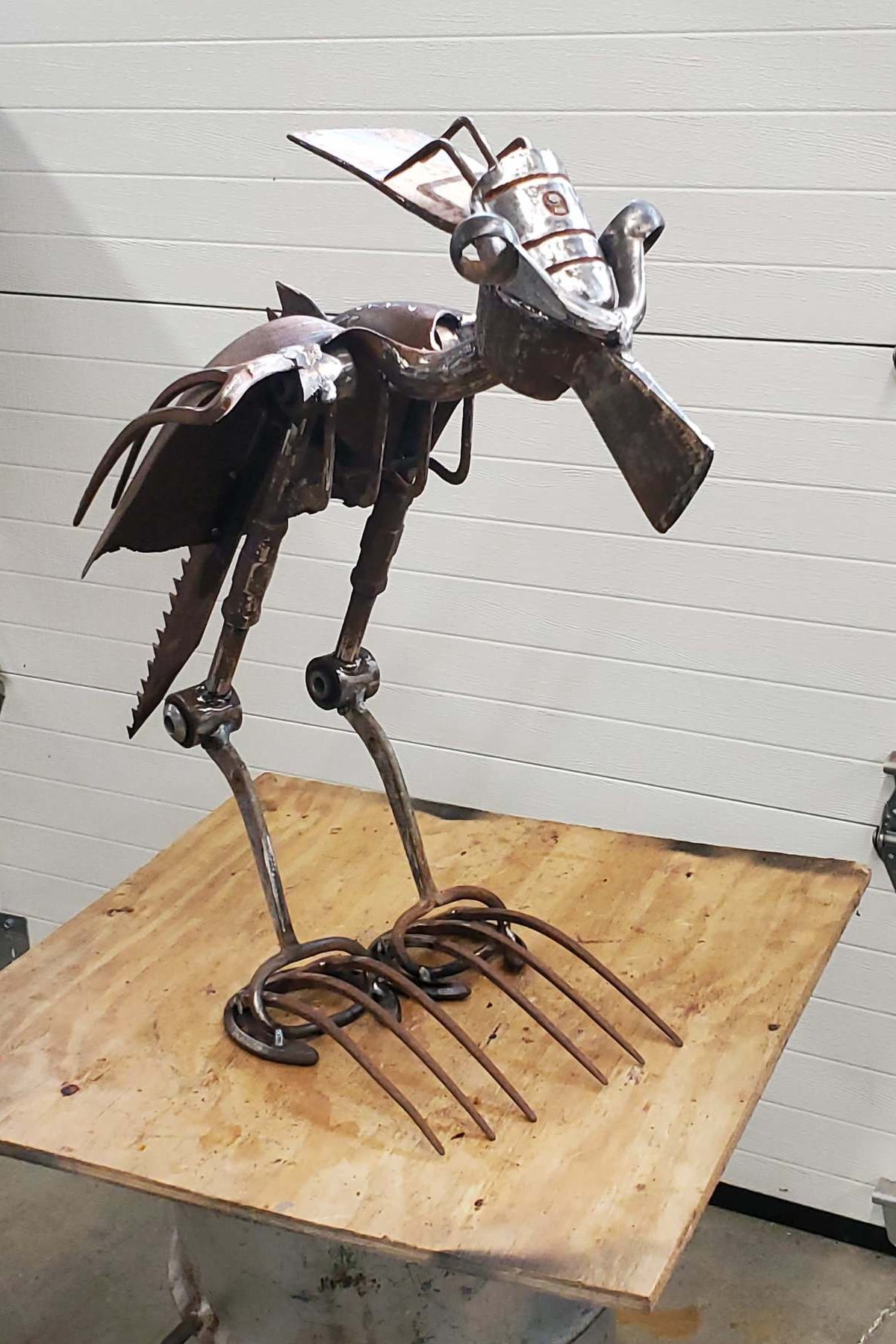 Metal sculpture of a bird with long legs and a beak, sitting on a wooden table. The sculpture is made from various metal scraps and parts, giving it a rustic and artistic appearance.
