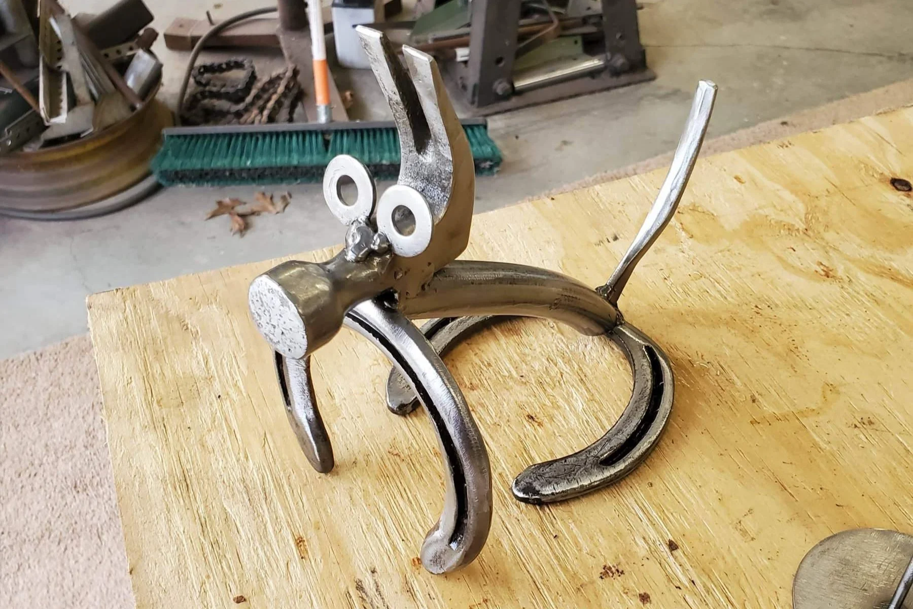 Metal sculpture resembling a cartoonish dog with googly eyes, elongated head, and curved legs, placed on a wooden surface in a workshop setting.
