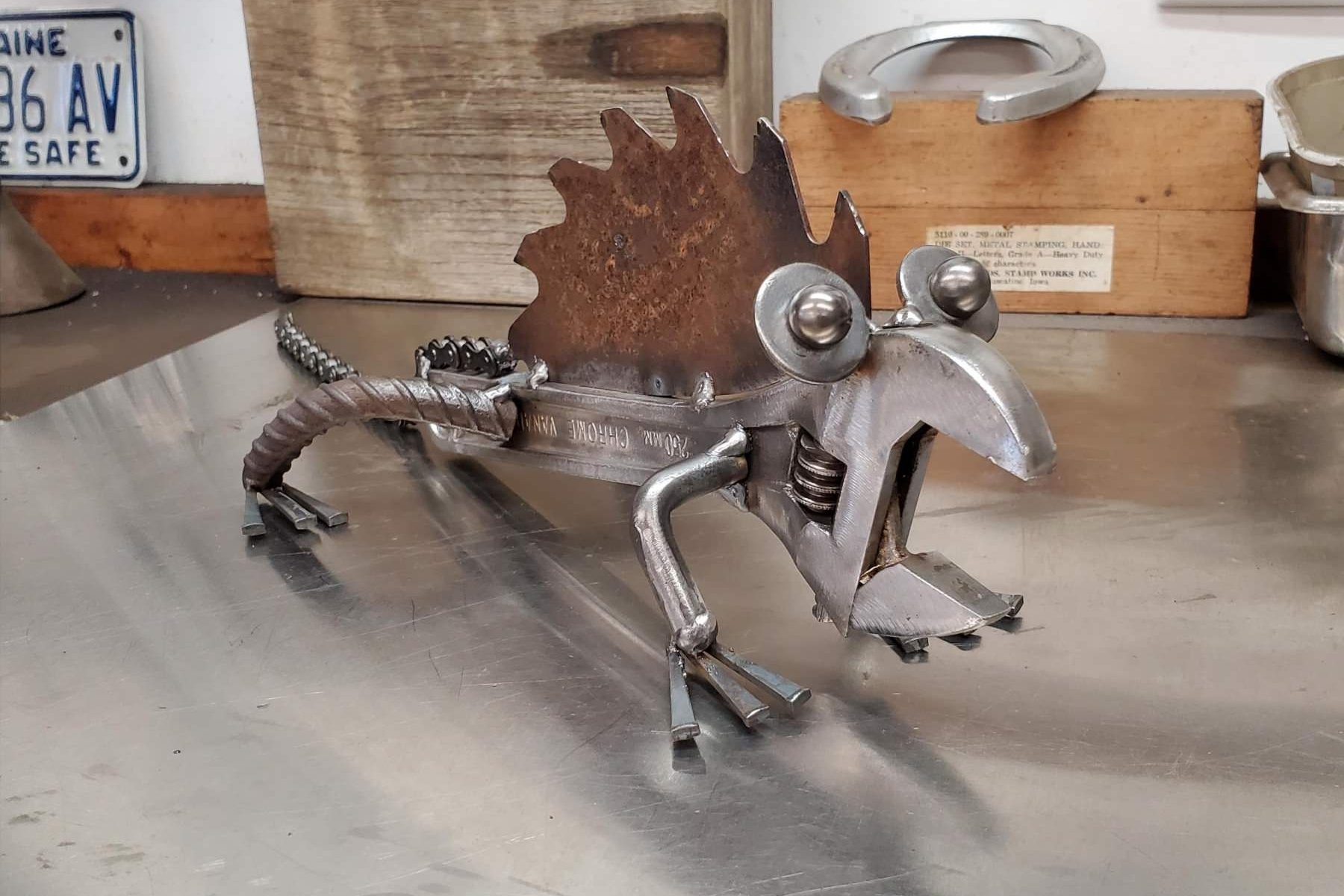 A sculpture of a lizard made from metal scraps, featuring a metal leaf as a tail, metal spheres as eyes, and various metal pieces forming the body and legs, placed on a metal surface with a wooden box and part of a license plate in the background.