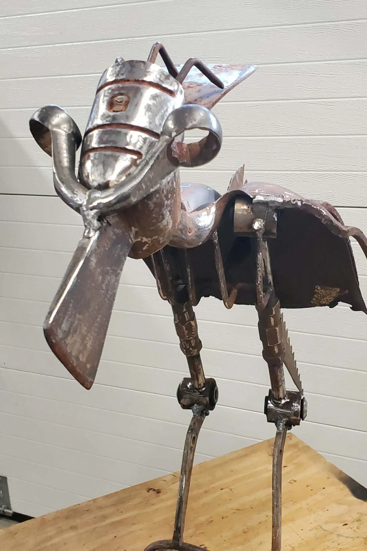 A metal sculpture of an animal made from various metal parts, including a coiled wire for the mouth, a pipe for the body, and rods for the legs, standing on a wooden surface against a light-colored wall.