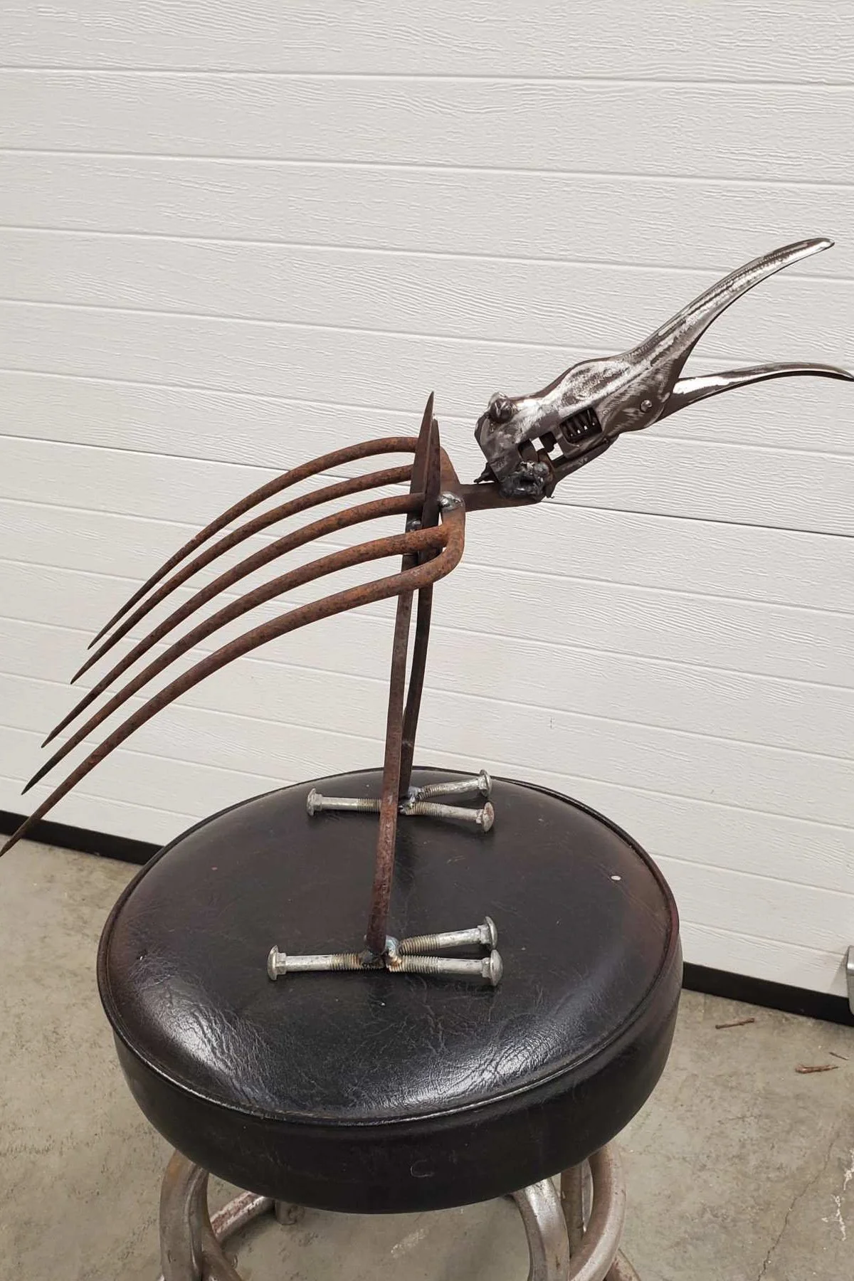 Metal sculpture of a bird with a sharp beak, composed of rusted metal parts, placed on a black cushioned seat with scattered screws underneath, set against a white paneled wall.