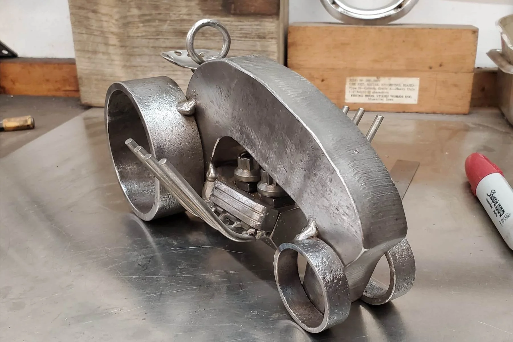 Art sculptures made from metal parts resembling a car, placed on a wooden table against a white paneled wall.