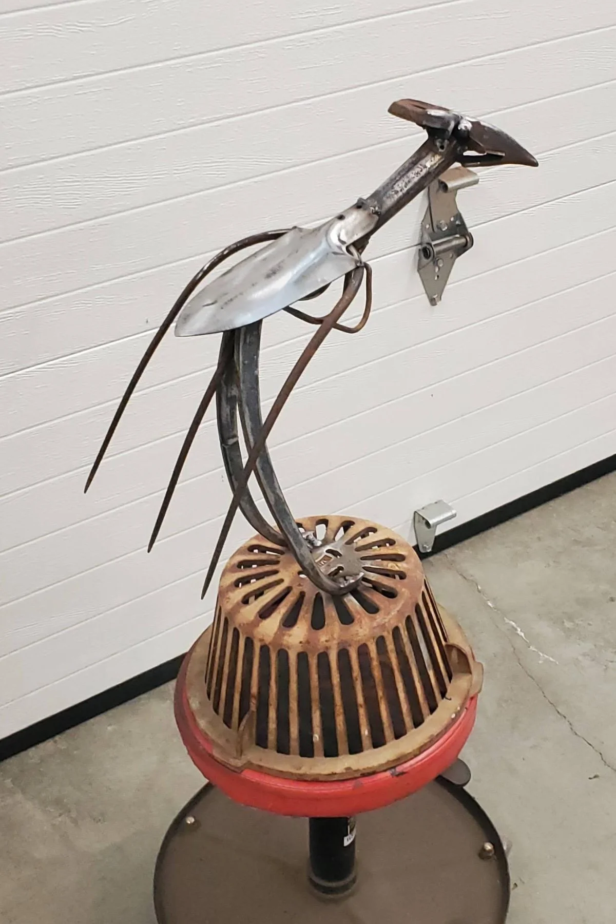 A sculpture made from scrap metal parts resembling a bird, mounted on a chair, against a white shiplap wall.