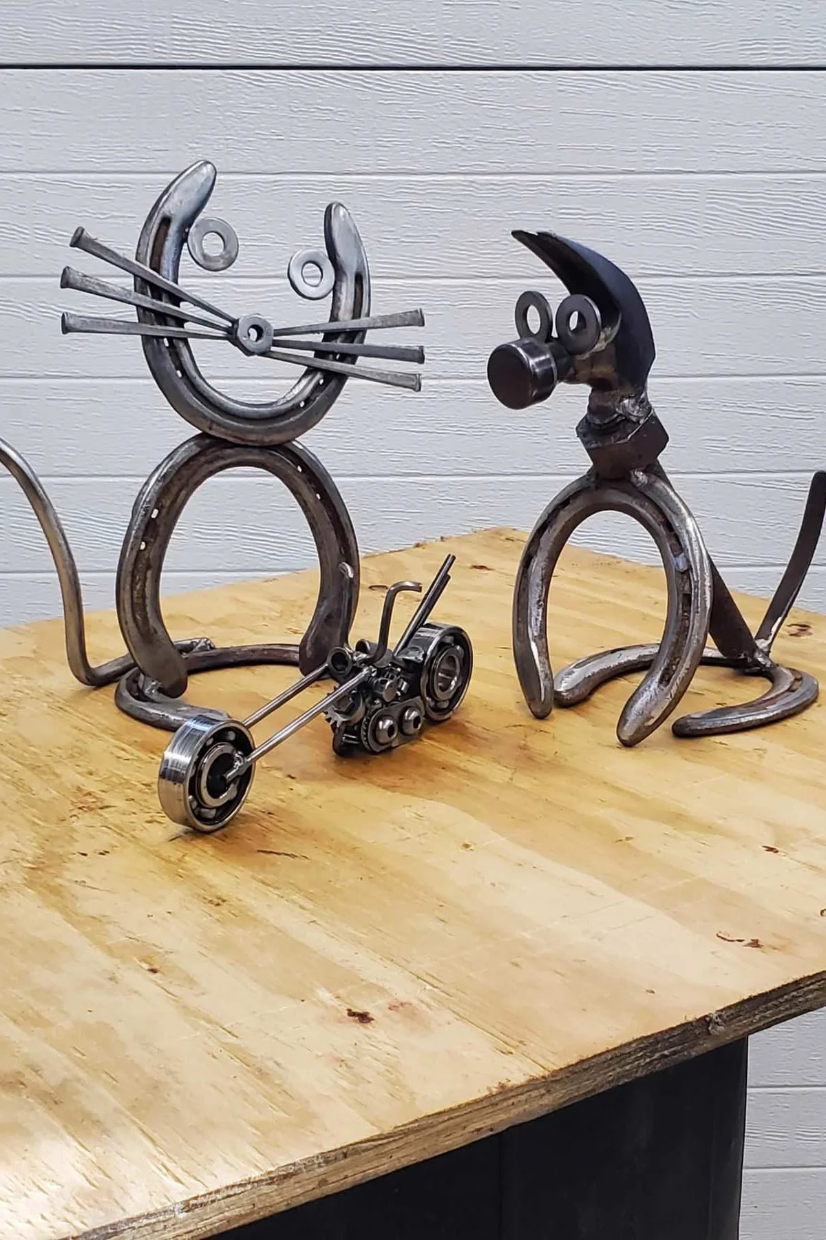 Art sculptures made from metal parts resembling animals, placed on a wooden table against a white paneled wall.