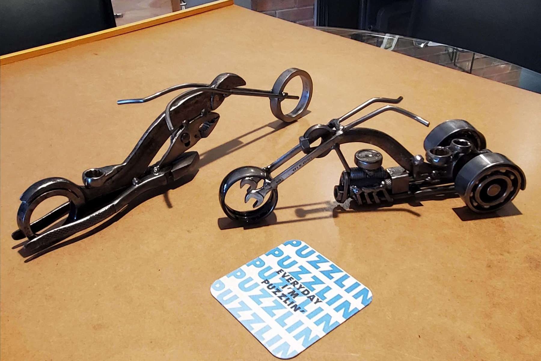 Three sculptures made of metal parts resembling motorcycles and a wrench, displayed on a wooden table.