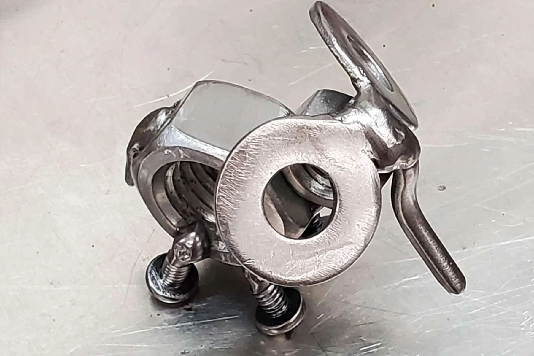 A small metallic elephant figure with a round body, two spring-like legs, a circular head with a large hollow center, and a small protruding arm, sitting on a metallic surface.