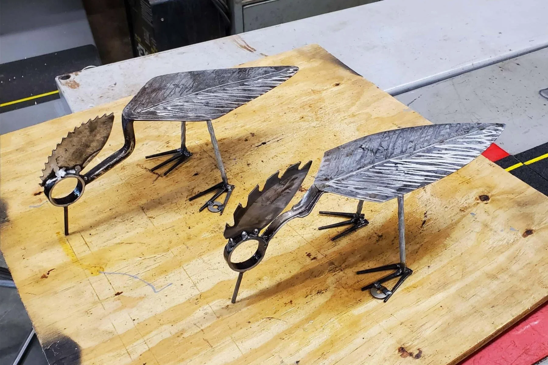 Two metal bird sculptures with leaf-shaped wings and circular heads with ring eyes, standing on a wooden workbench.