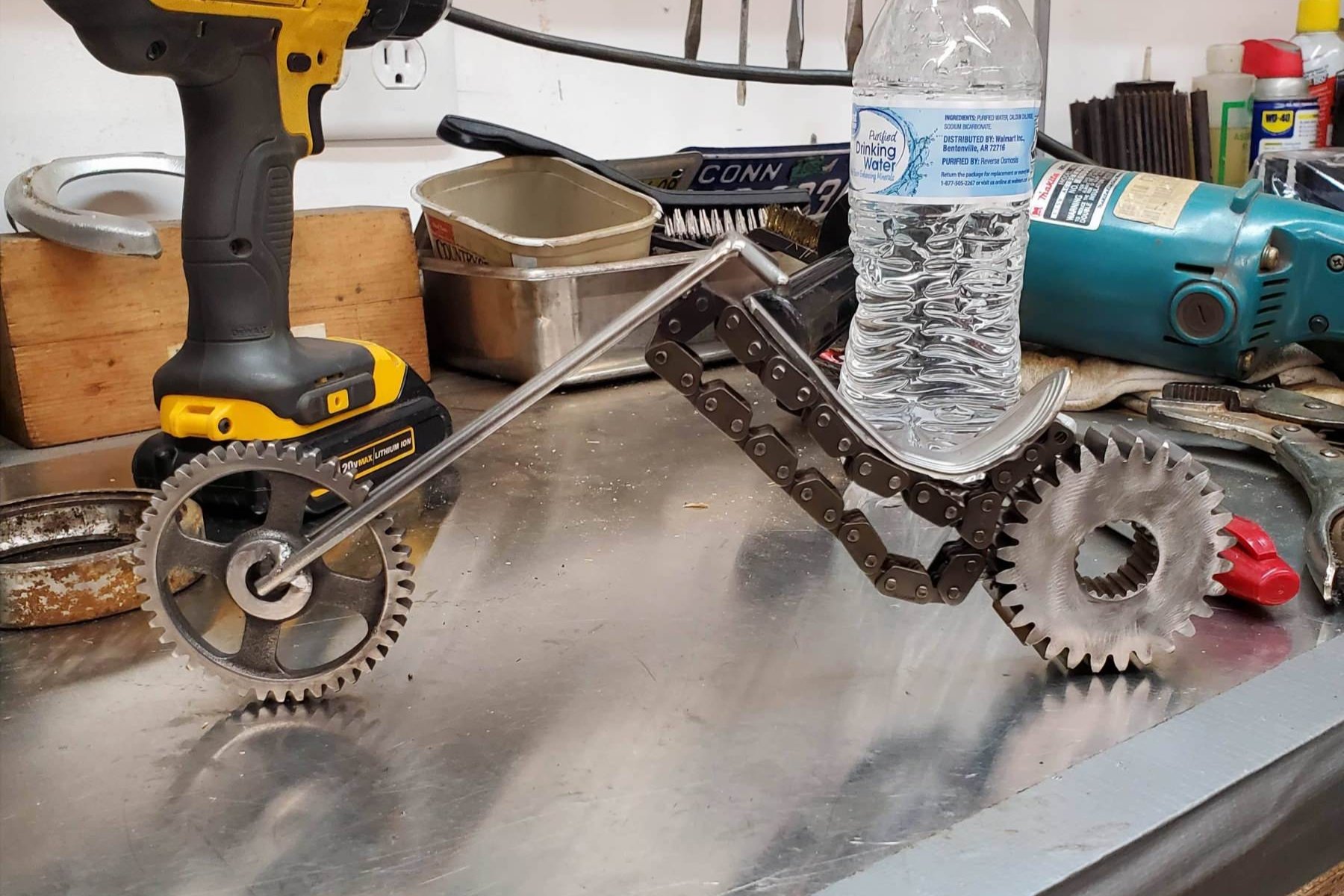 A miniature robotic vehicle made from gears, chains, and metal rods on a workshop workbench with tools and water bottle in the background.