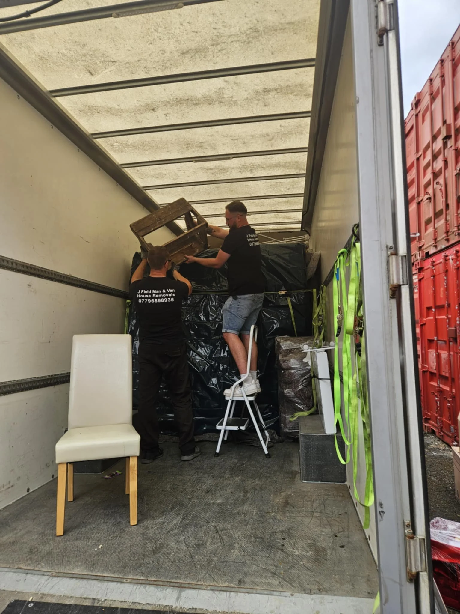 Two men working inside an open moving truck, one is standing on a step stool and holding a small wooden table, the other is assisting, with a white chair in the foreground and stacked large black plastic bags and boxes in the truck.