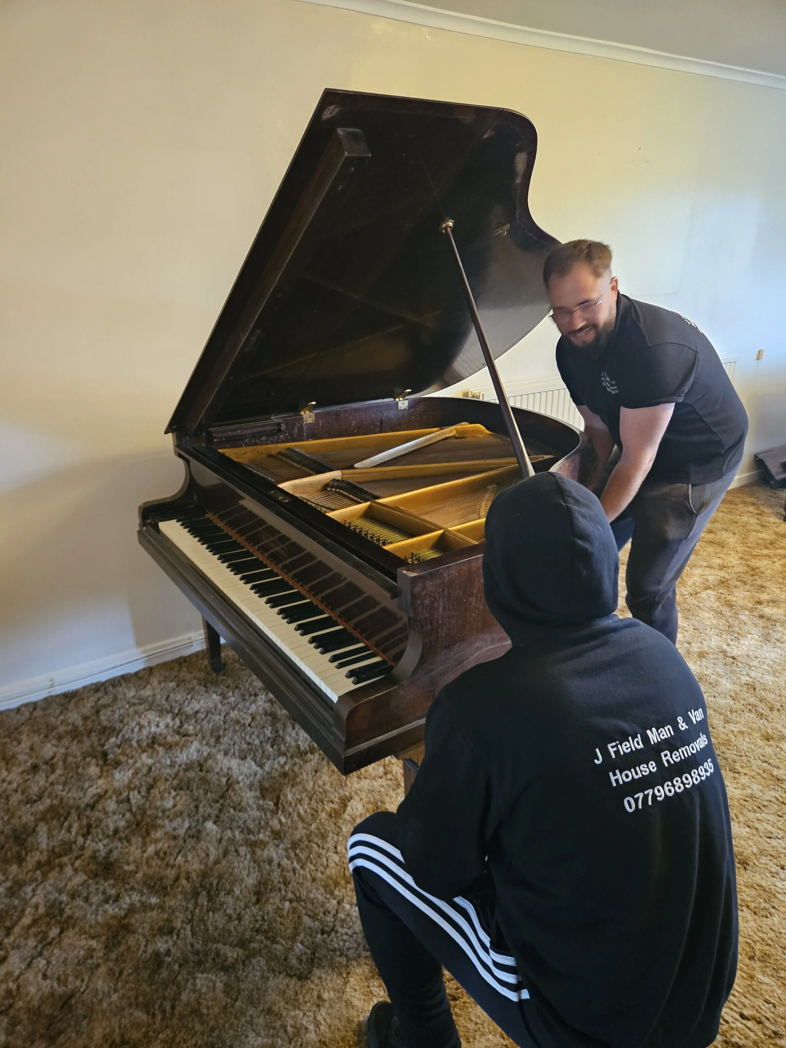 Two men are working on moving a grand piano inside a room with beige carpet and light-colored walls. One man is leaning over the piano, and the other is crouching with his back to the camera, wearing a black hoodie with the company logo on the back.