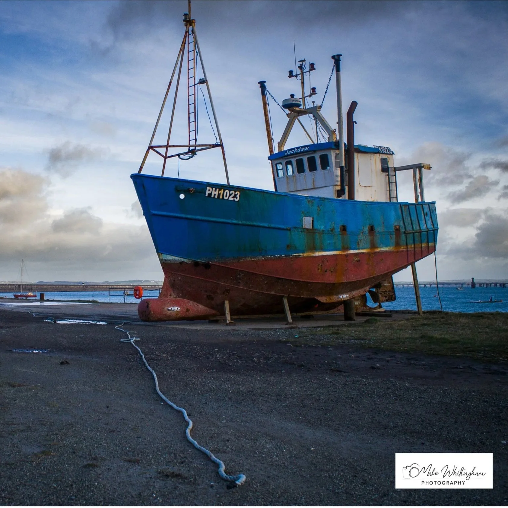 A large, weathered blue and red fishing boat resting on land near the water, with a cloudy sky in the background.
