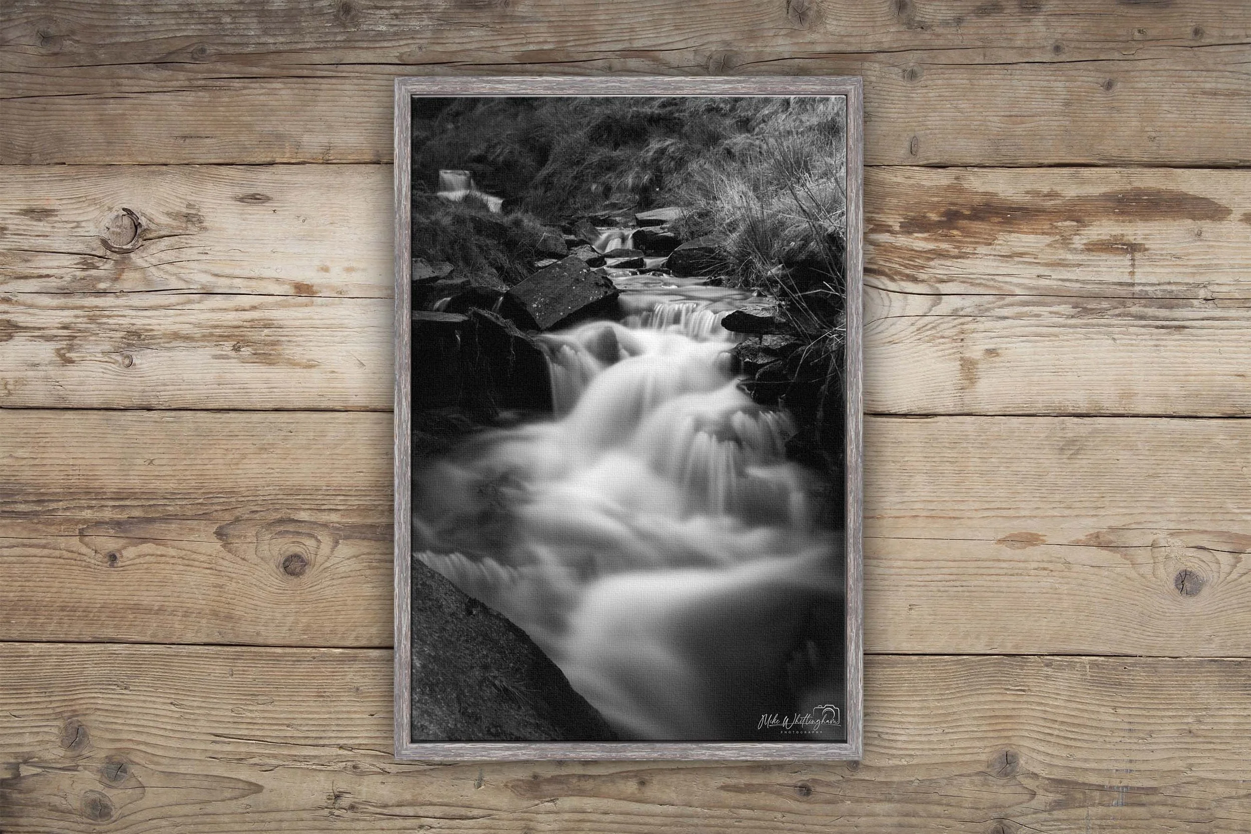 Black and white photograph of a flowing creek with rocks and trees, signed by the artist in the corner, framed and hung on a wooden wall.