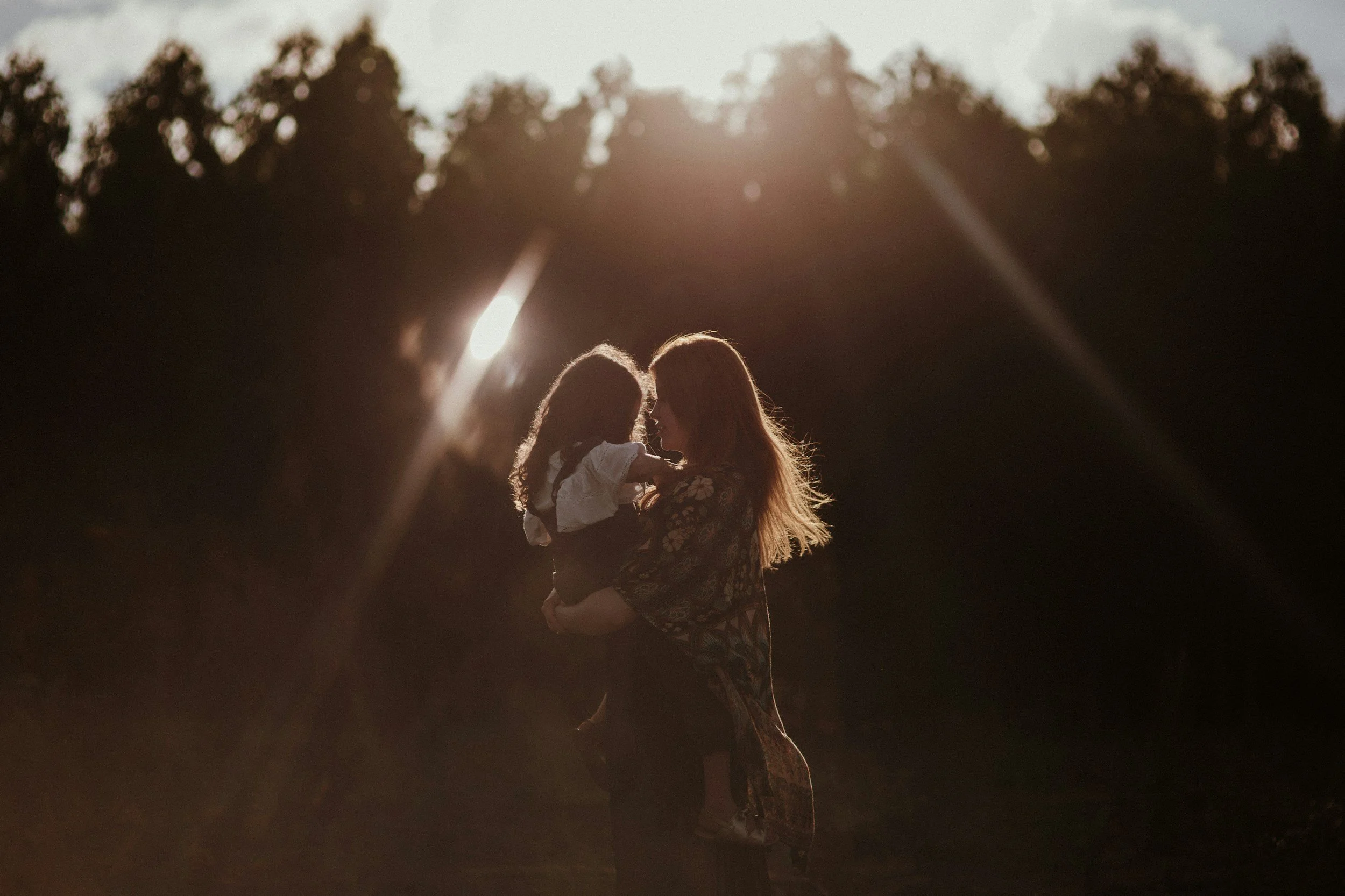 A woman holding a child in an outdoor setting during sunset, with greenery and trees in the background.