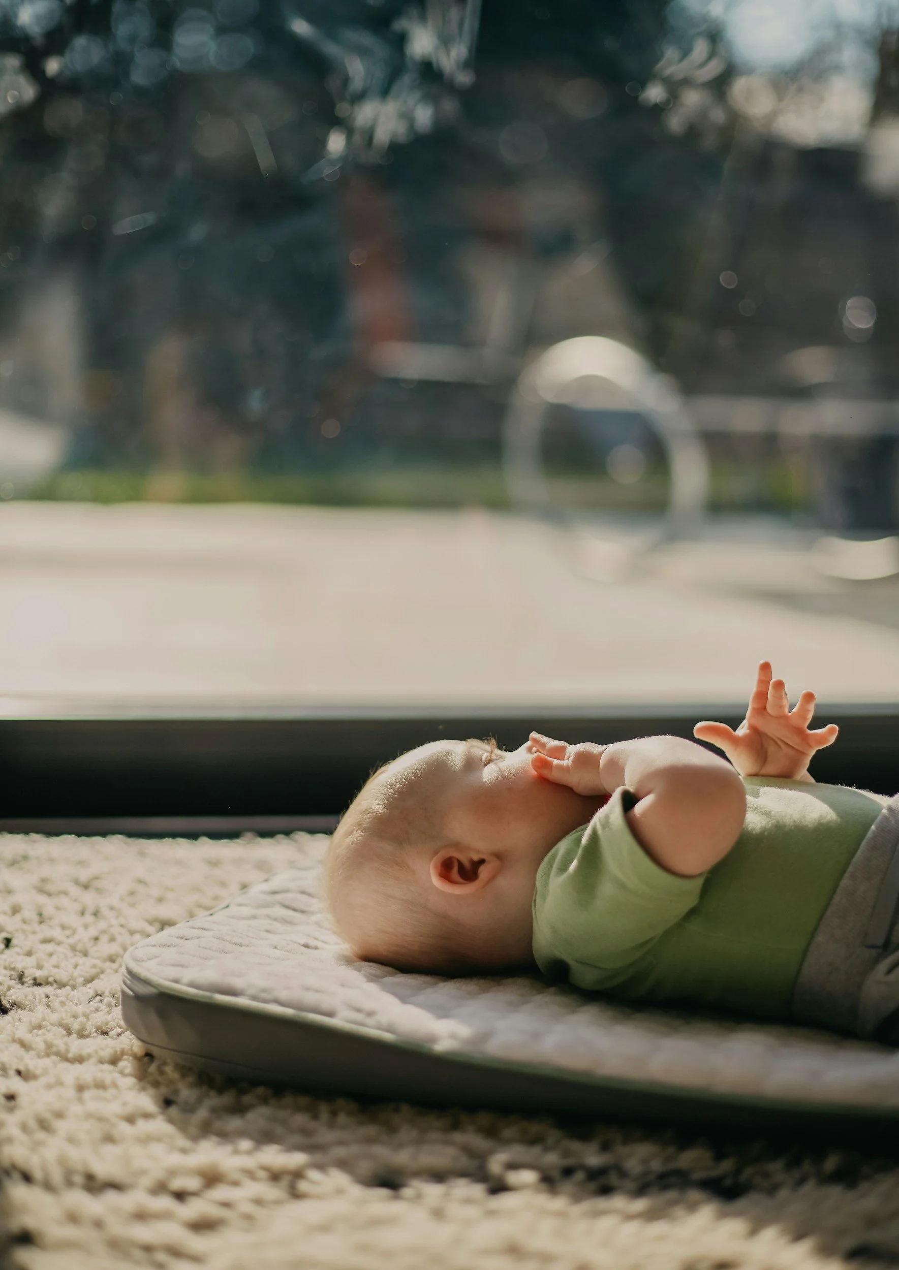 Young baby rests on mattress on rug looking at sunny sky through living room window.