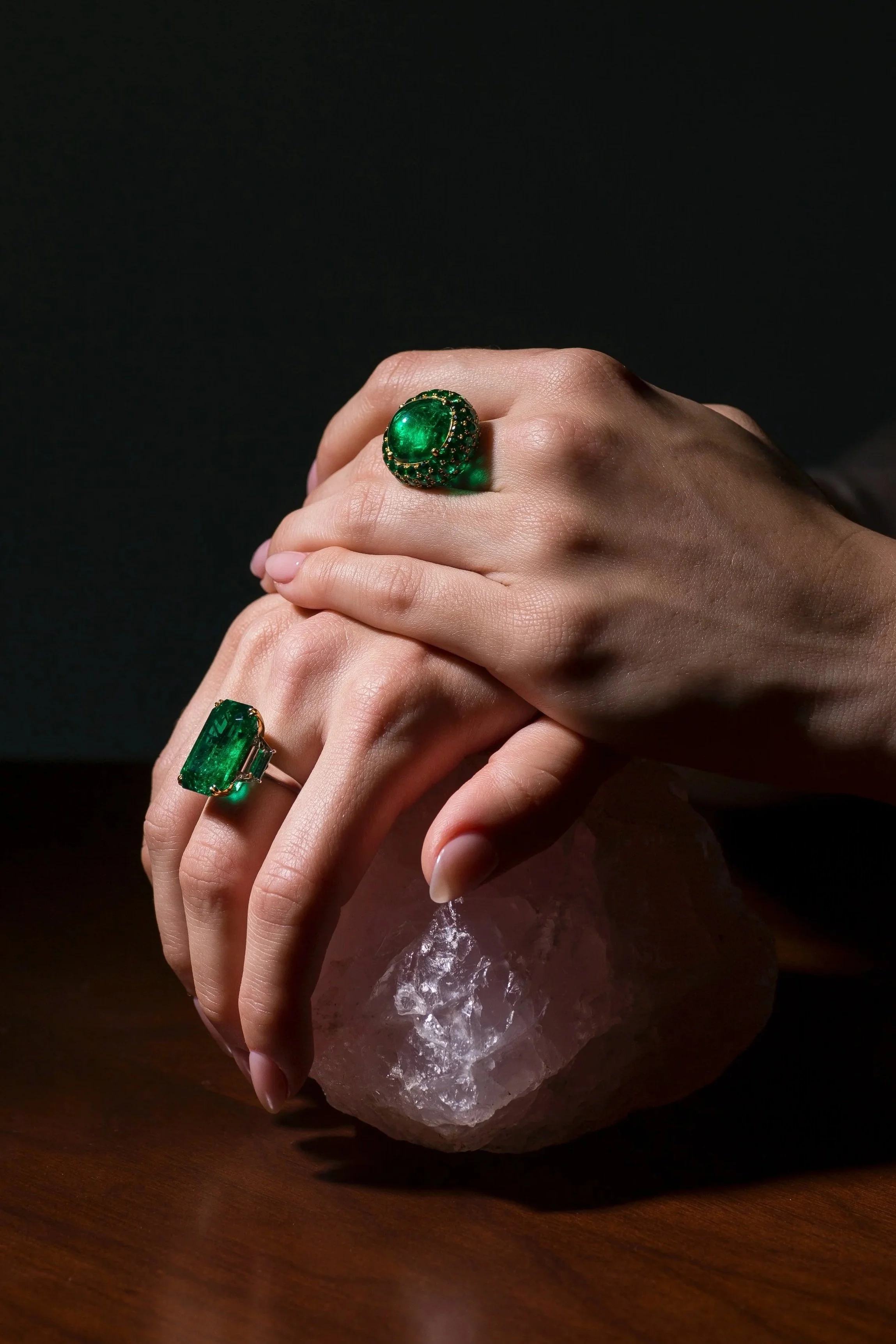Two hands with large Colombian emerald rings holding a rough pink stone against a dark background.