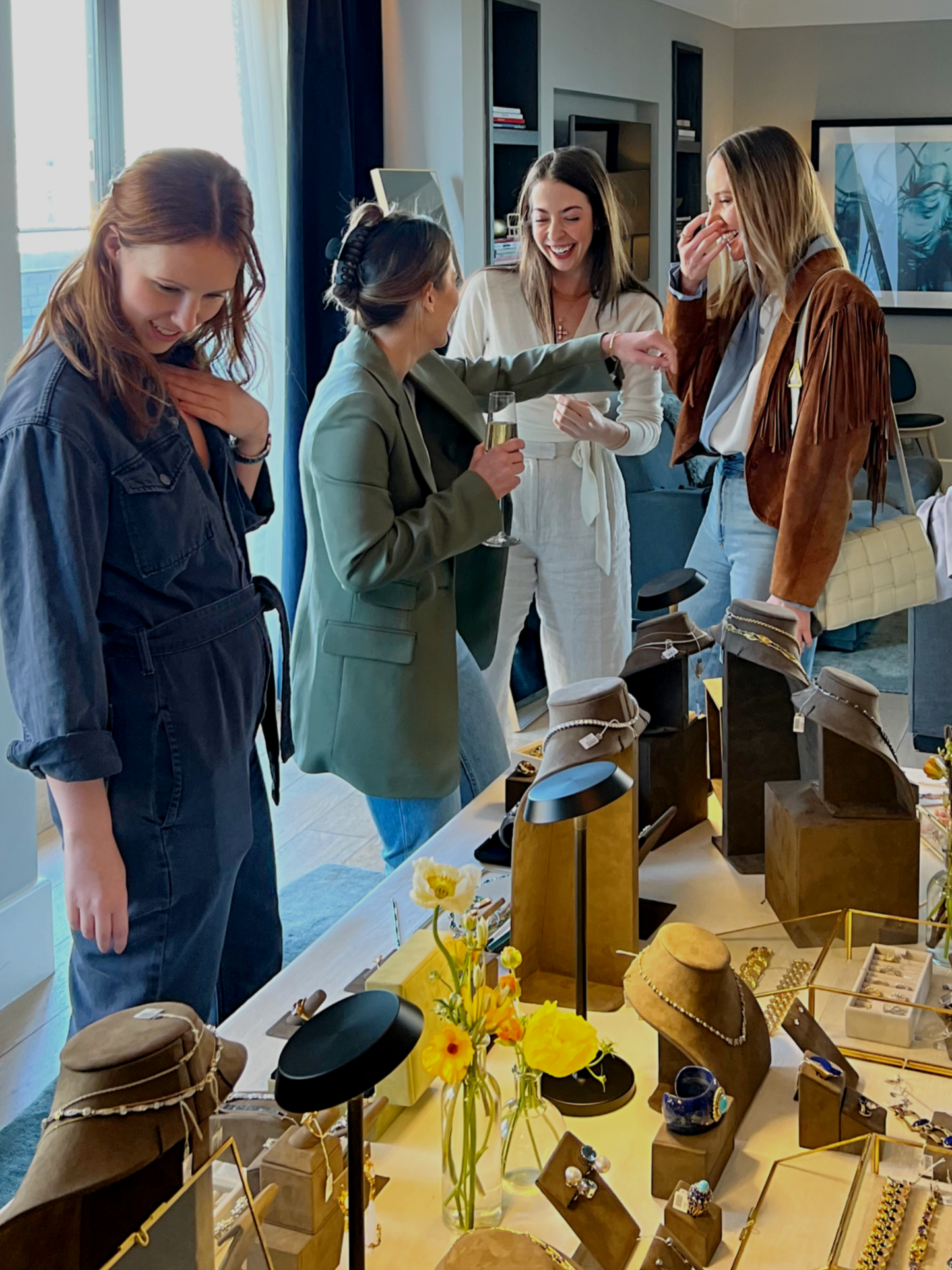 Four women look at jewelry displayed on a table inside a showroom in Soho in downtown Manhattan. One woman is holding a glass of champagne. They are smiling and appear to be enjoying shopping or browsing jewelry.