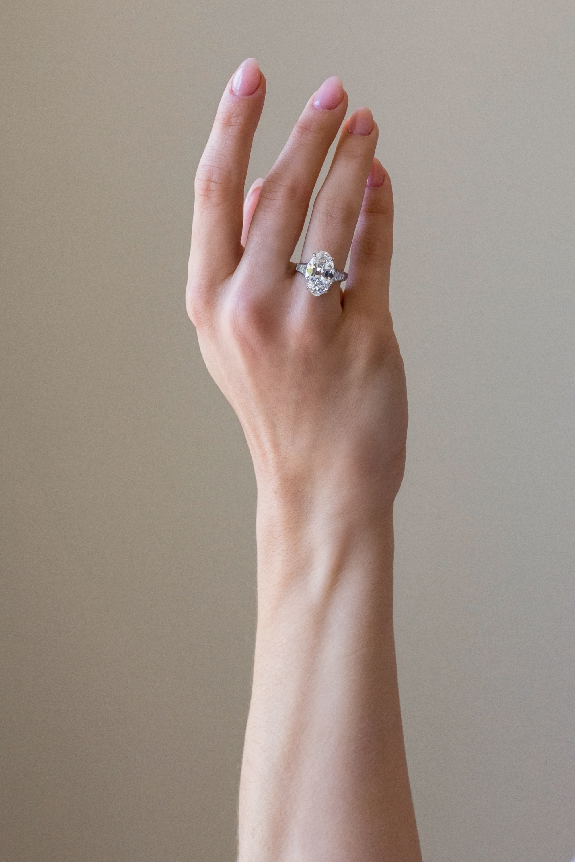 A close-up of a woman's hand raised with nails painted in soft pink, wearing a large diamond engagement ring on her ring finger, against a blurred beige background.