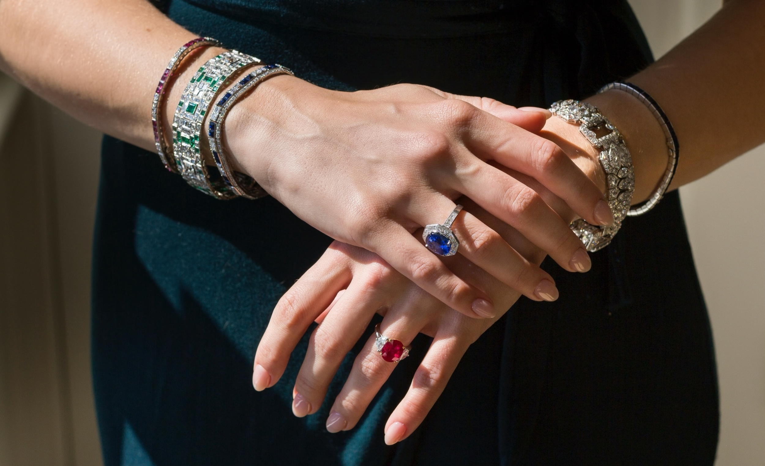 Close-up of a person's hands with vintage Art Deco jewelry, including rings and bracelets featuring blue sapphire, emerald, and rubies, with one hand resting on top of the other.