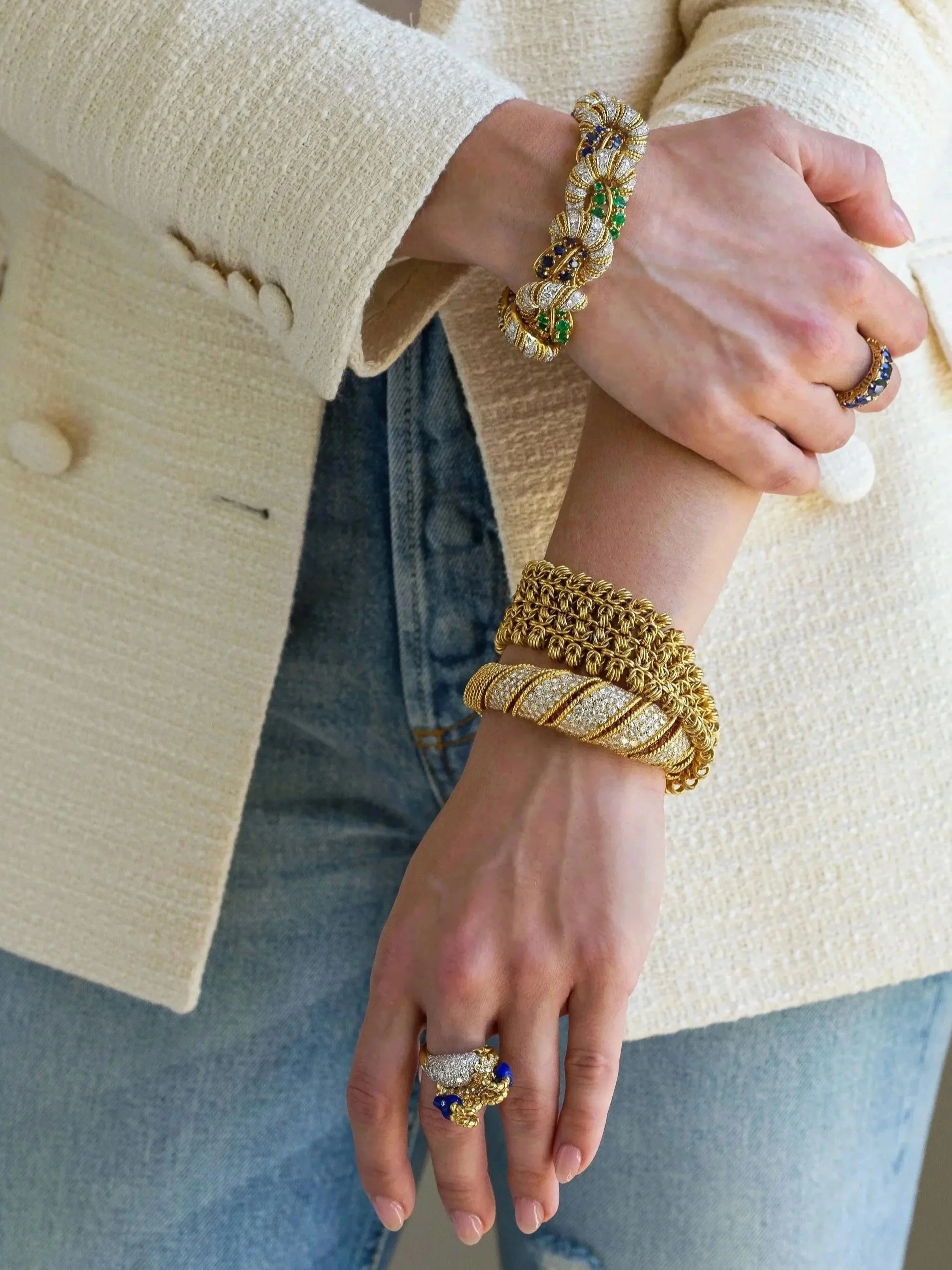 Close-up of a person wearing multiple gold jewelry pieces, including bracelets, rings, and a chain, with a beige textured blazer and light blue jeans.