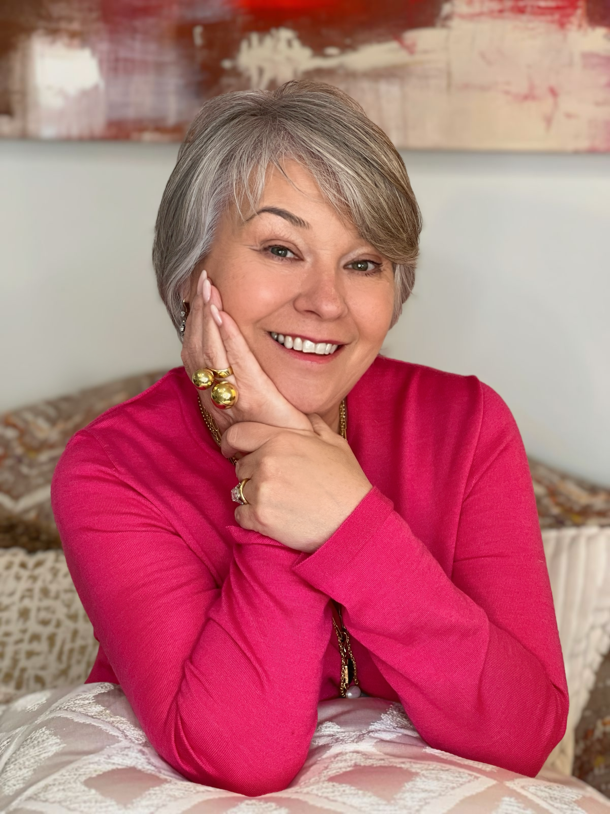 Donna Vock A smiling middle-aged woman with short gray hair, wearing a pink top and gold jewelry, sitting on a bed with a beige patterned blanket, in front of an abstract painting.