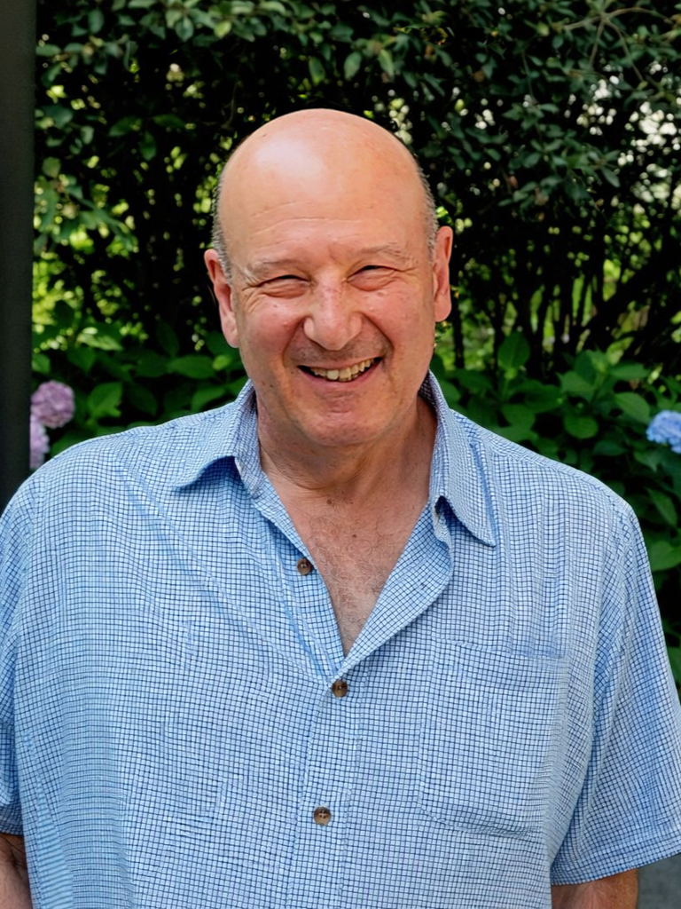 A smiling middle-aged man with a bald head, wearing a light blue checkered shirt, standing outdoors in front of green shrubbery and flowers.