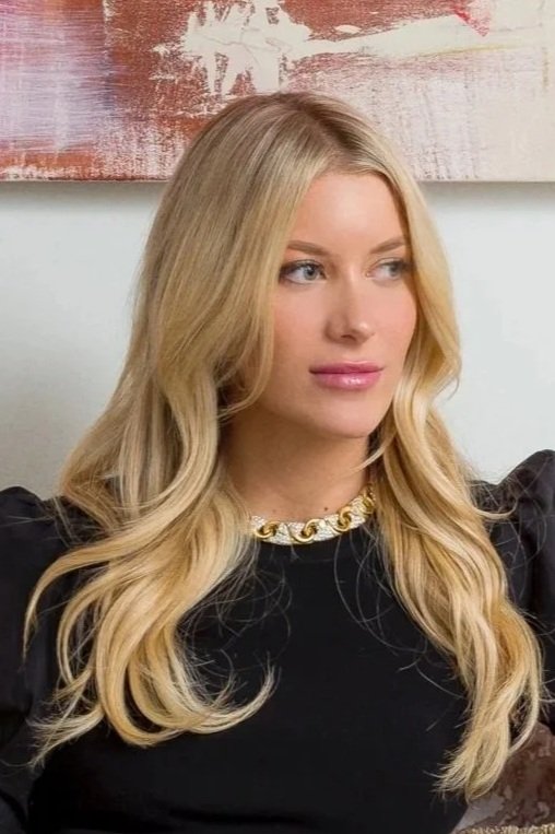 A blonde woman with wavy hair wearing a black top and a gold chain necklace, sitting indoors with an abstract painting in the background.