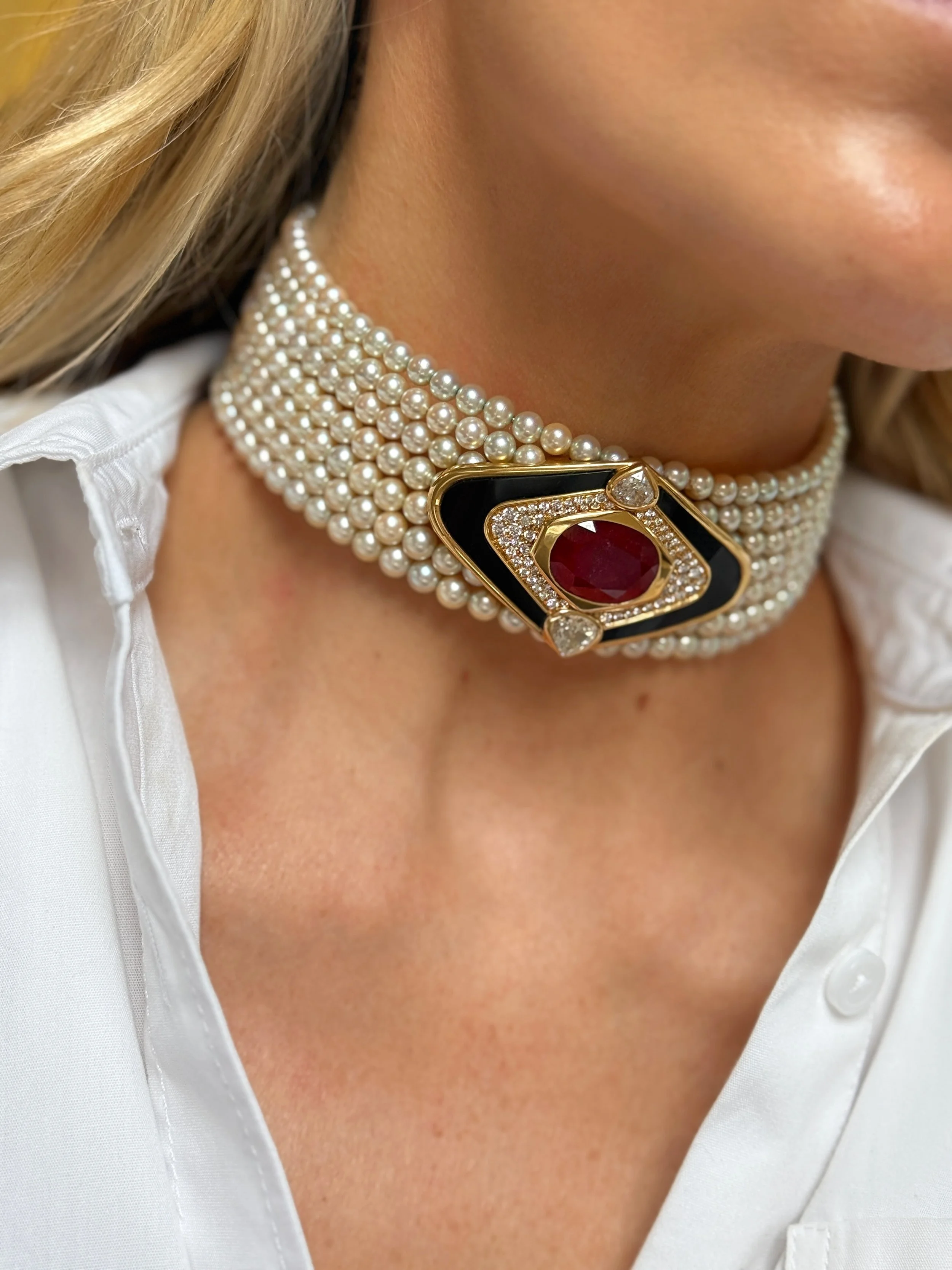 A woman wearing a vintage Marina B multi-strand pearl choker with a large, ornate gold and gemstone clasp featuring diamonds, a red gemstone, and black accents, paired with a white collared shirt.