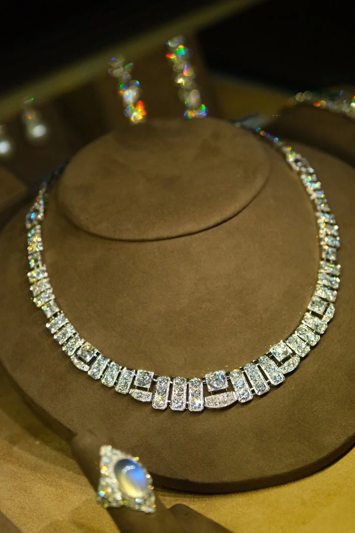 An Art deco diamond necklace displayed on a brown jewelry bust, with matching earrings and a ring also adorned with diamonds.